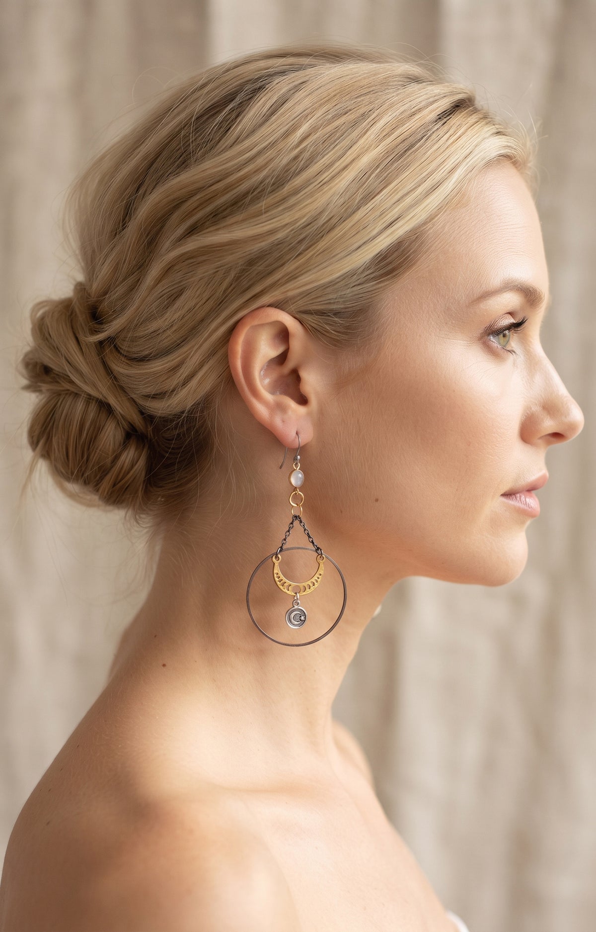 Chandelier Moonphase Earrings by Circle of Heather worn by a model with blonde hair in a low twisted updo, opalescent stone above gold rings, an oxidized hoop and a gold moon phase crescent with a silver medallion.