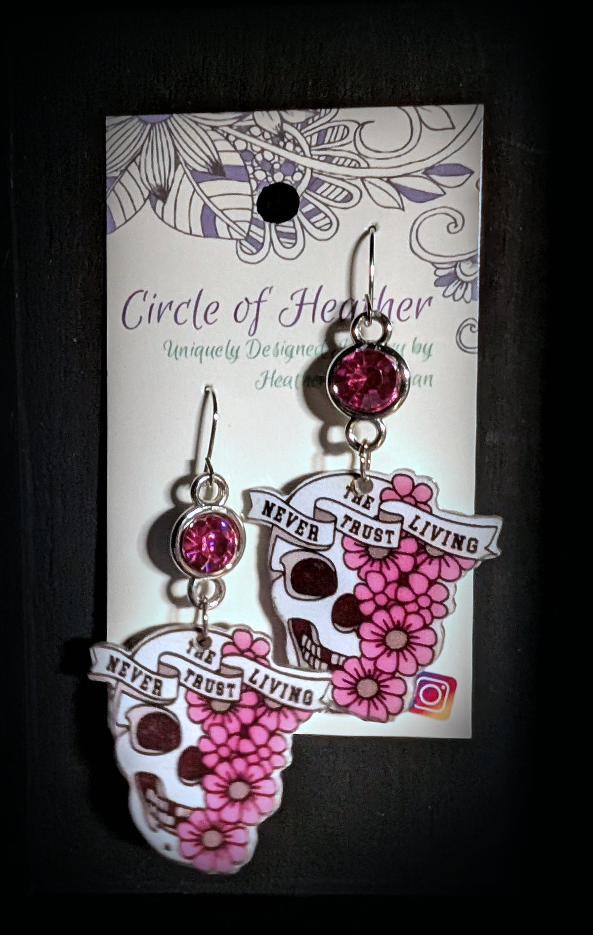 A pair of dangling earrings displayed on a branded card against a dark background. The earrings feature skull and flower motifs with pink gemstones and the words 'NEVER TRUST THE LIVING.