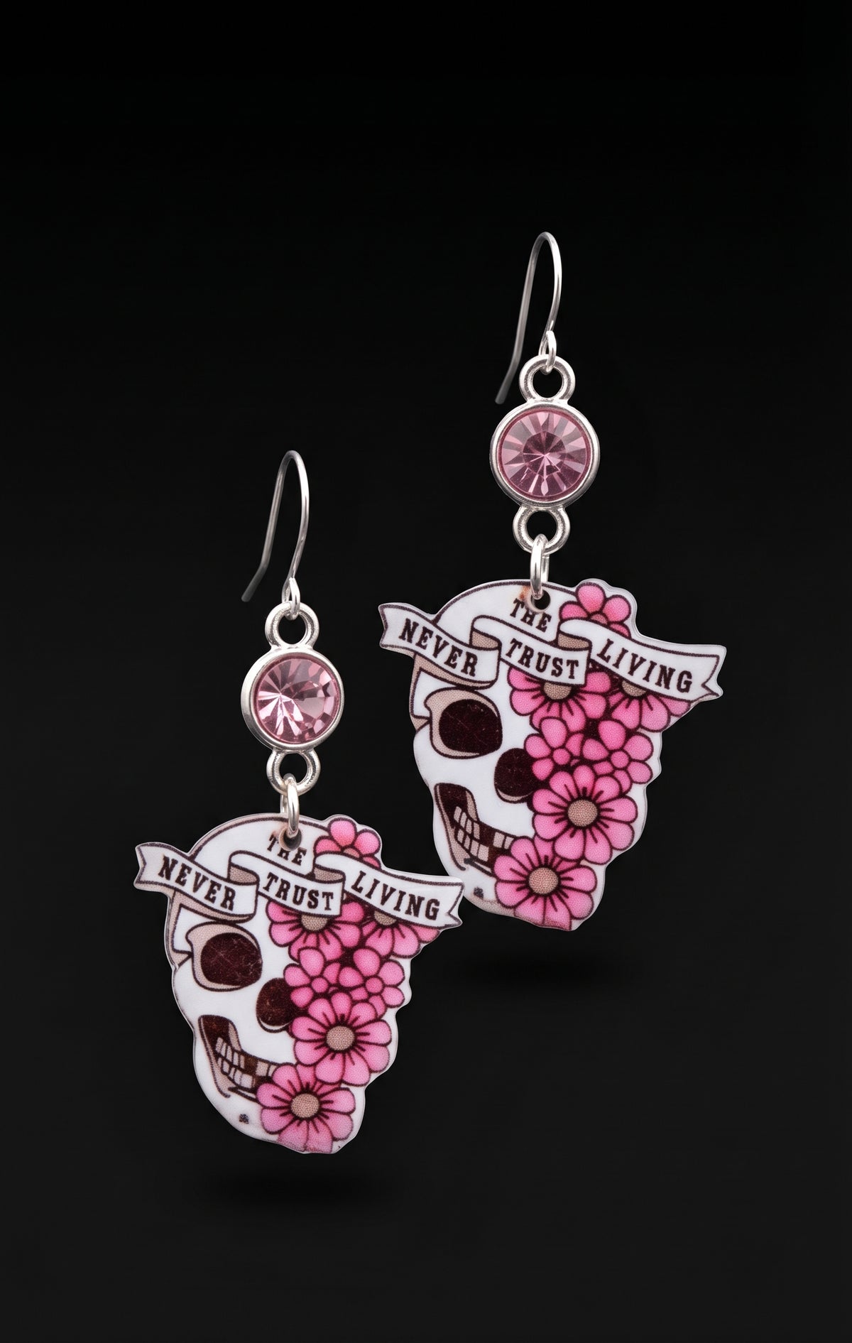 A pair of dangling earrings on a black background. Each earring has a silver fishhook, a round pink gem, and a sugar-skull charm decorated with pink flowers and banners reading 'NEVER TRUST THE LIVING'.