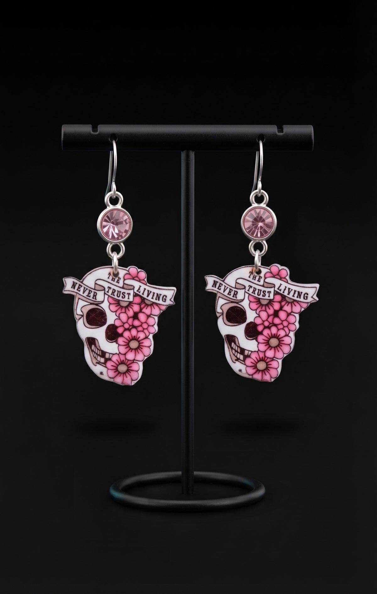 A pair of dangling earrings displayed on a black stand against a black background. Each earring has a silver hook, a round pink gem, and a sugar skull charm decorated with pink flowers and a banner that reads 'NEVER TRUST THE LIVING".