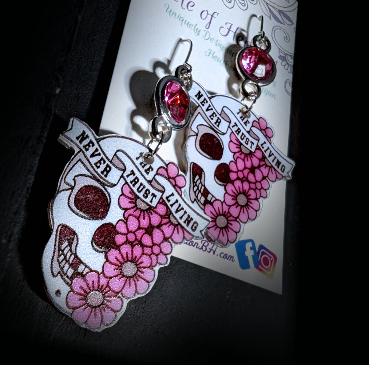 A pair of gothic sugar skull dangle earrings displayed on their branded backing card. The earrings feature a white sugar skull design decorated with pink flowers and a silver banner that reads 'NEVER TRUST THE LIVING'.