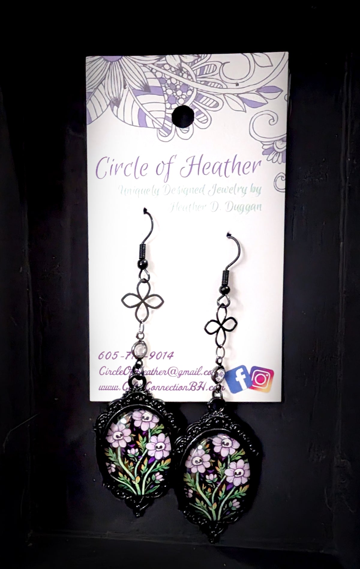 Ornamental Framed Skully Flower Earrings by Circle of Heather on the original branded card, black filigree oval frames with pale purple skull-faced flowers under glass, hanging from black chain with a quatrefoil link