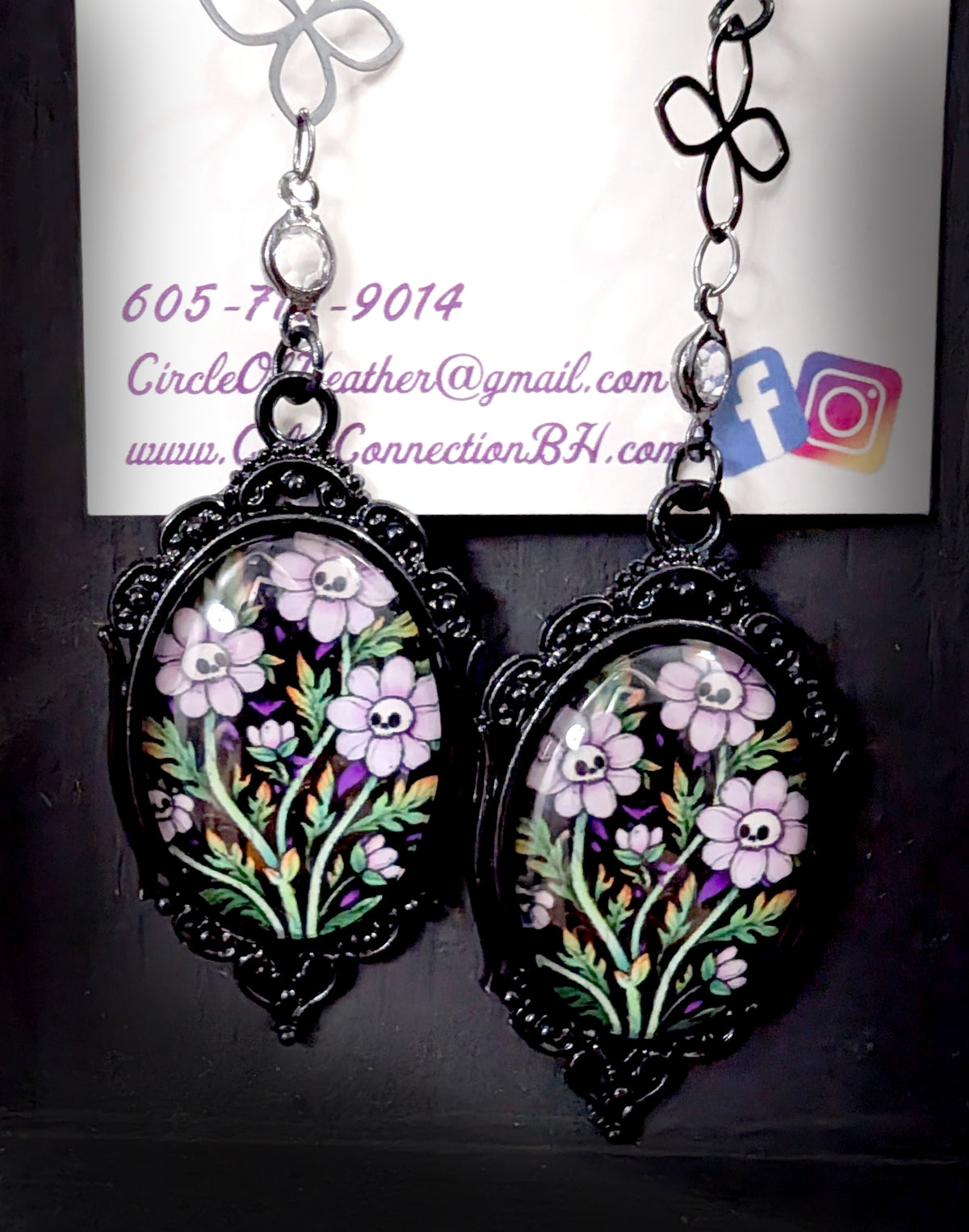 Ornamental Framed Skully Flower Earrings by Circle of Heather, black filigree frames with hand-illustrated lavender skull flower artwork, hanging beside Circle of Heather packaging card
