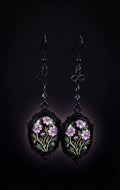 Ornamental Framed Skully Flower Earrings by Circle of Heather, black filigree oval frames with pale purple skull-faced flowers under glass, hanging from black chain with a quatrefoil link and dark crystal bead