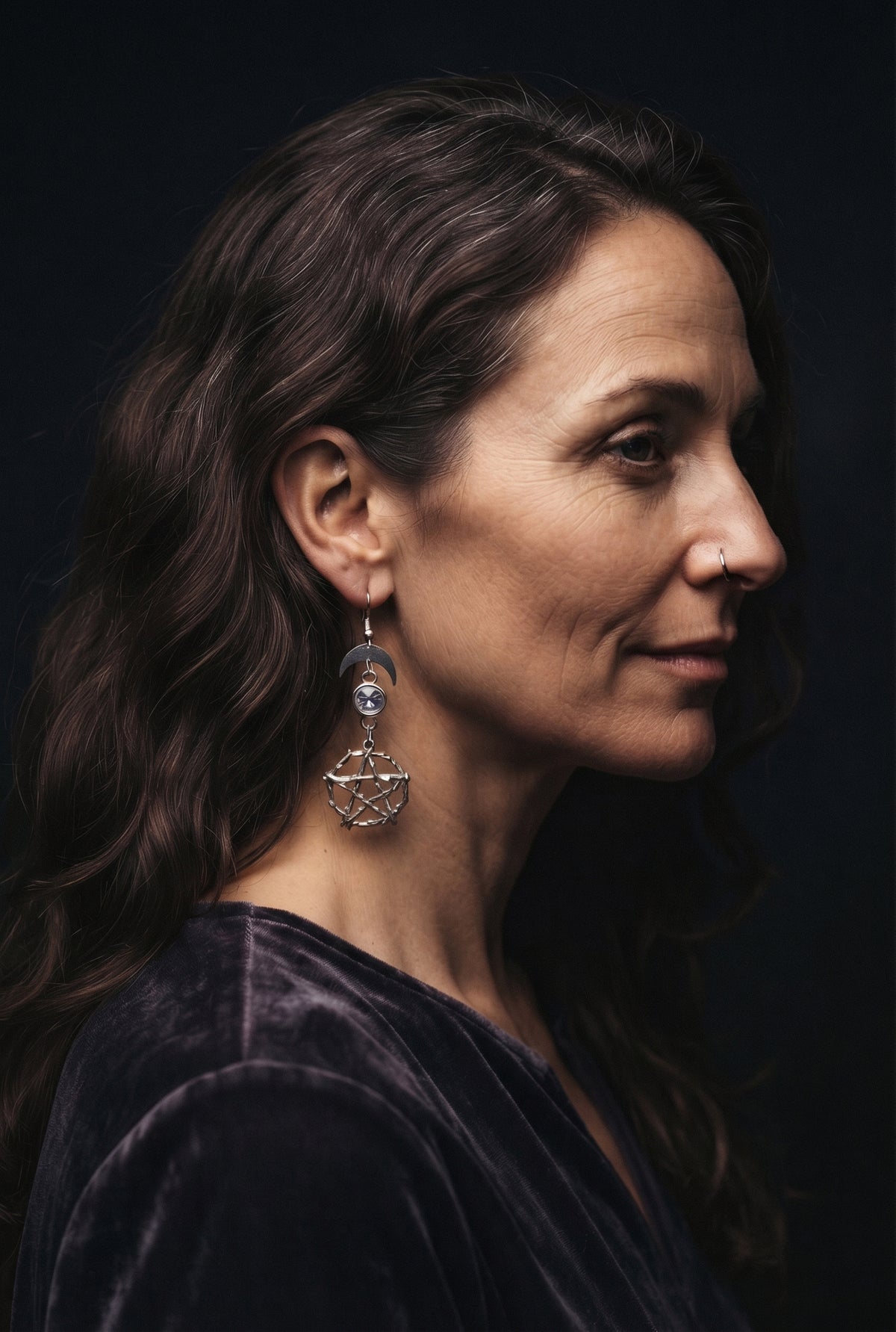 Pentacle Lunar Blue Blush Earrings by Circle of Heather worn by a model with long wavy brown hair and a nose ring, silver crescent moon above a pale blue stone and a twig pentacle, shown with a purple velvet top.