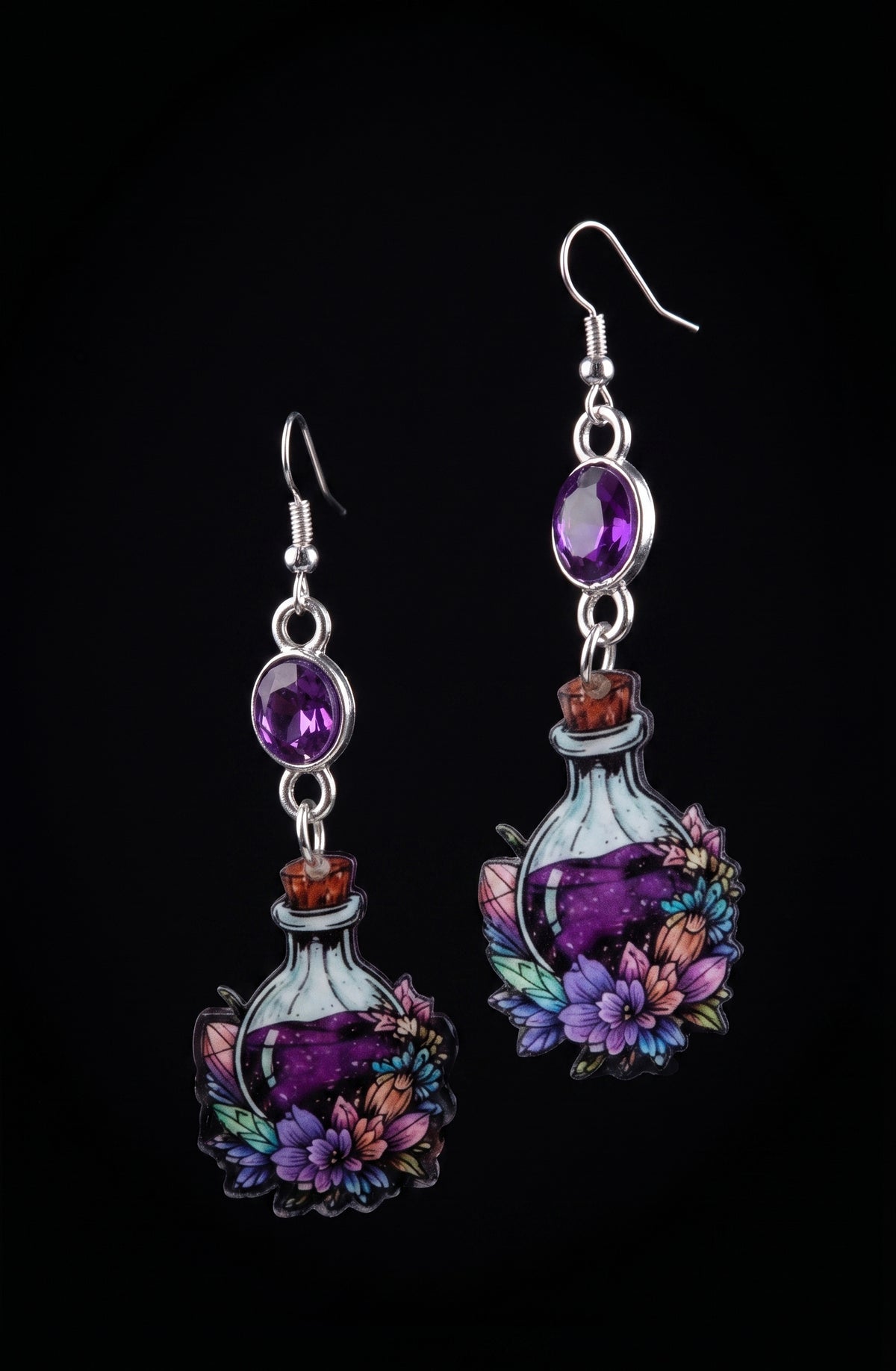 A pair of dangling earrings shaped like small illustrated potion bottles with purple liquid, silver hooks, oval purple stones, and colorful floral details, shown on a black background.