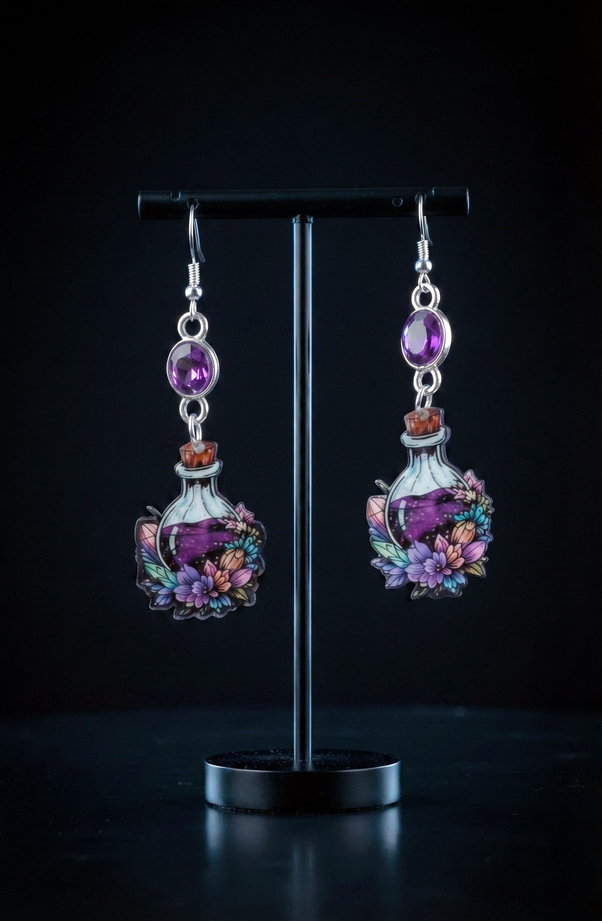 A pair of dangling earrings displayed on a black stand against a dark background. Each earring has a silver hook and oval purple stone, with a small bottle charm featuring a purple liquid design and colorful floral details.