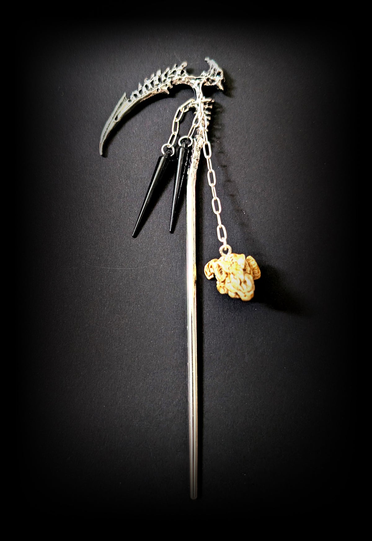 Isolated product shot of the silver spine-scythe hair pick against a black background, featuring two short chains with glossy black spikes and a longer chain dangling a weathered bone-colored ram skull charm.