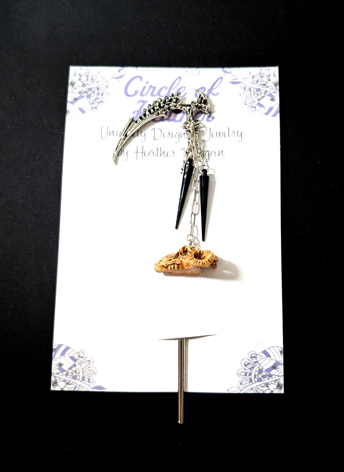 The silver spine-scythe hair pick attached to a white backing card featuring a decorative purple mandala pattern and text that reads "Circle of Heather, Uniquely Designed Jewelry by Heather Duggan."