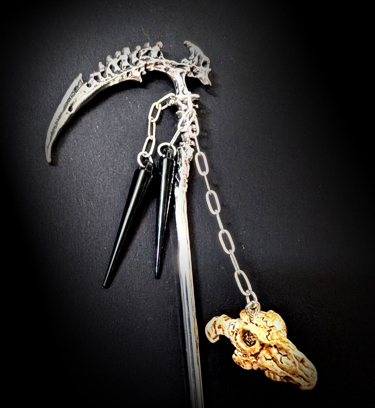 Close-up product shot against a black background highlighting the vertebra-cast blade with a spine ridge, the segmented spine shaft, and the dangling black spikes and ram skull charm.