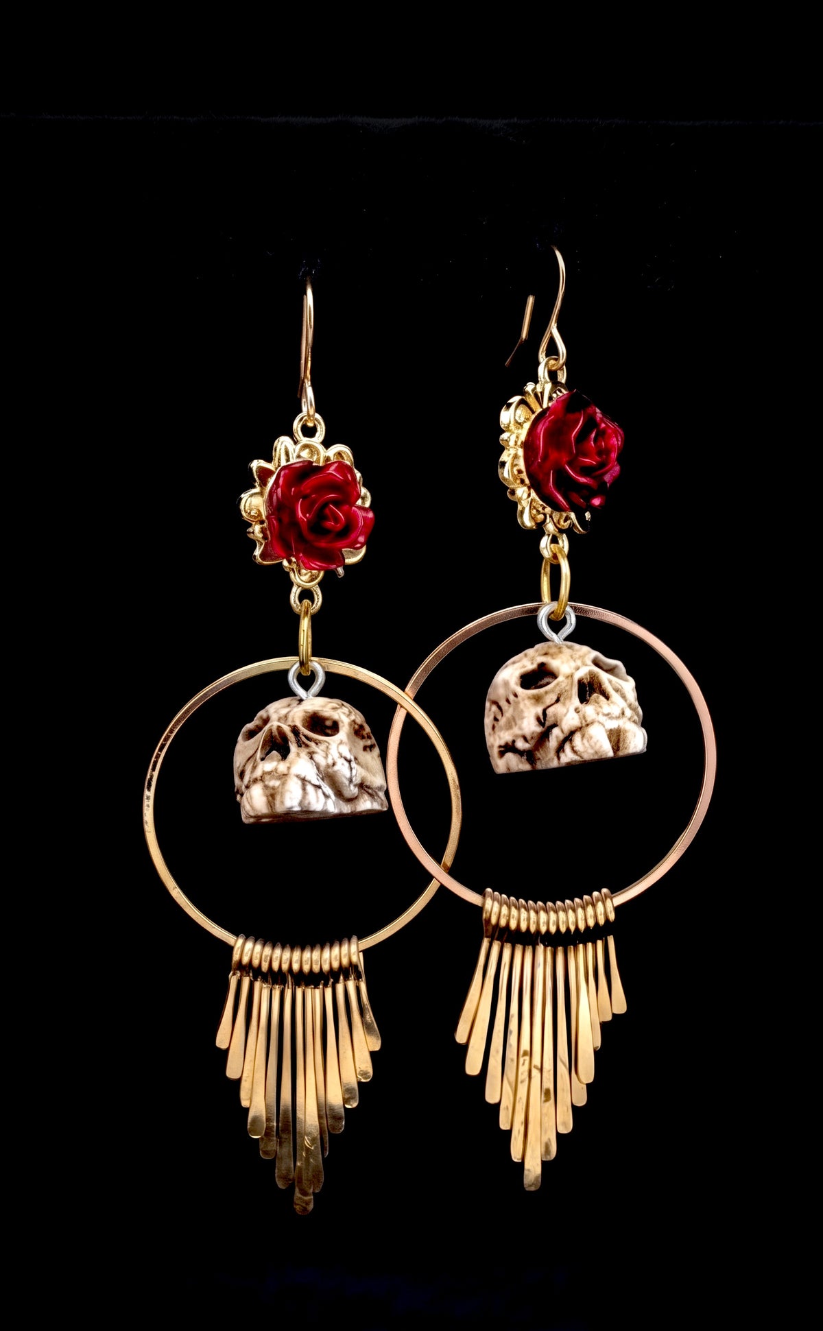 A pair of gothic-style gold dangle earrings featuring red roses, skull charms, and fan-shaped fringe, set against a solid black background.