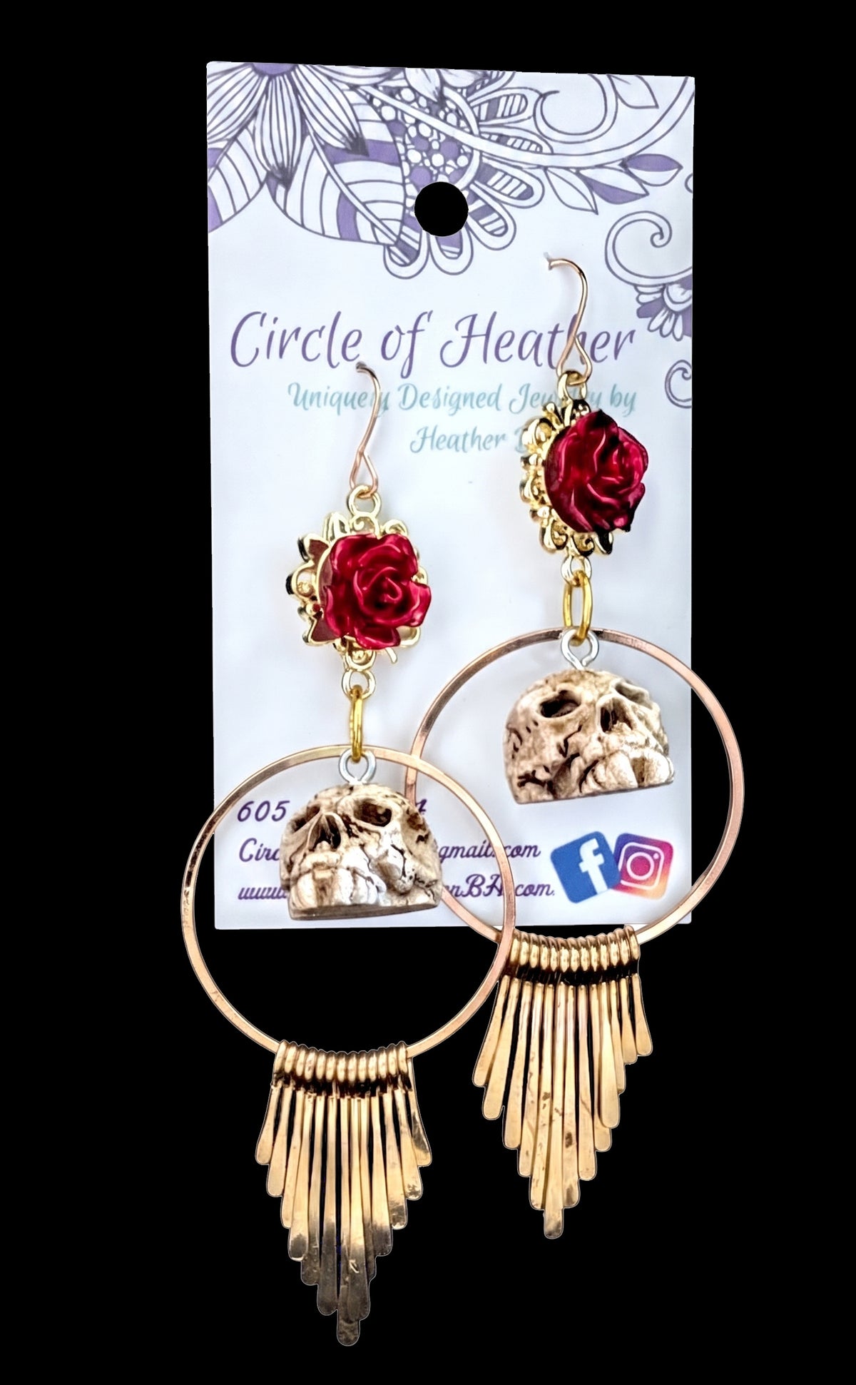 A pair of gothic-style dangle earrings displayed on their branded white backing card against a solid black background. The earrings feature red rose charms, skull pendants, and gold-tone circular hoops with fringe tassels.