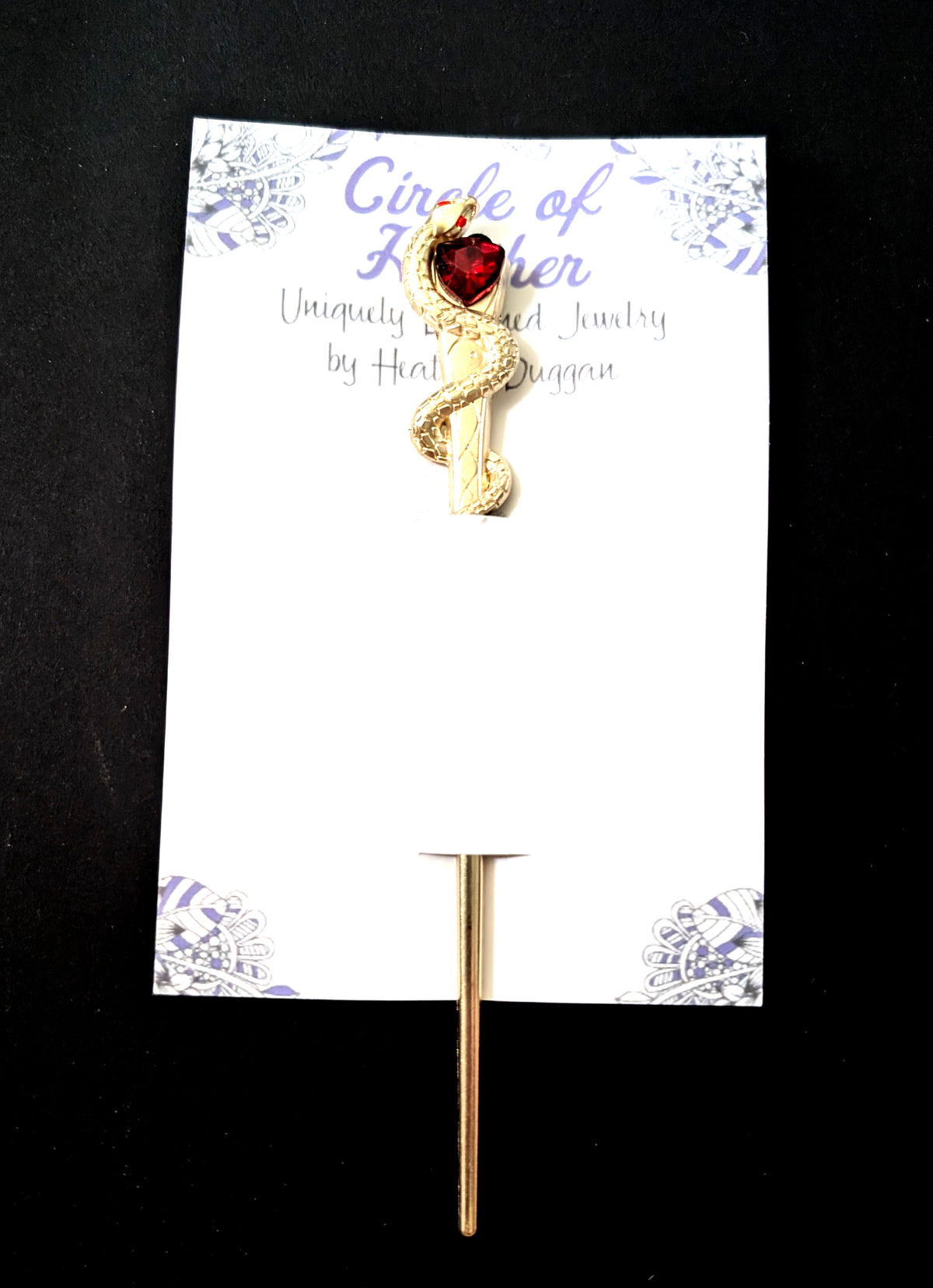 The gold-tone snake hair pick attached to a white backing card featuring a decorative purple mandala pattern and text that reads "Circle of Heather, Uniquely Designed Jewelry by Heather Duggan."
