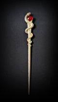 Isolated product shot of the slim gold-tone hair pick against a black background, featuring a scaled snake coiled around the upper shaft and a faceted red heart stone held at its head.