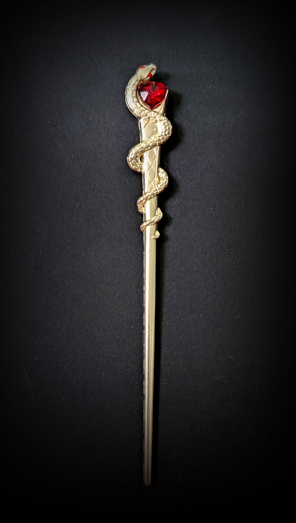 Isolated product shot of the slim gold-tone hair pick against a black background, featuring a scaled snake coiled around the upper shaft and a faceted red heart stone held at its head.