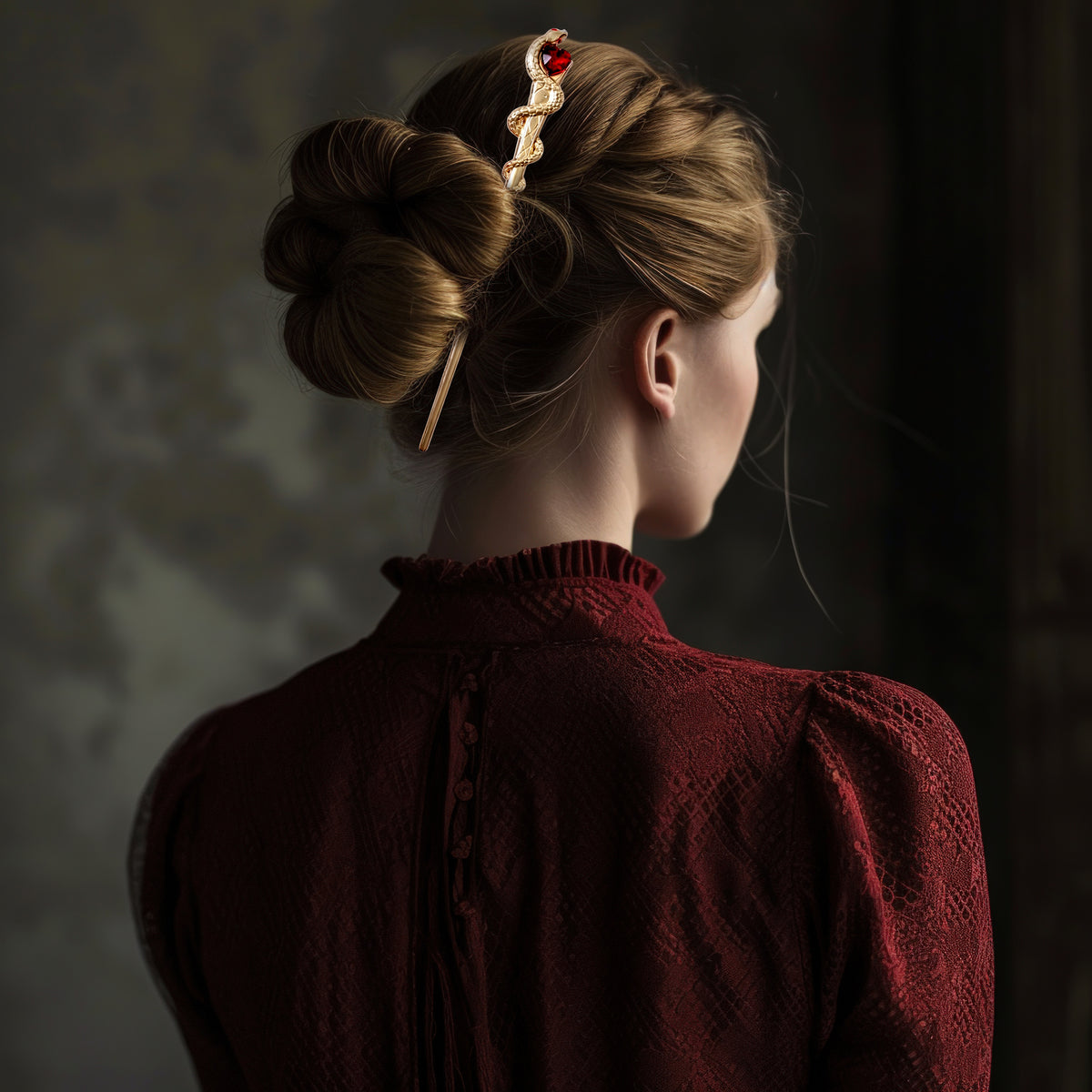 Side profile of a model with brown hair styled in a low bun secured by the gold snake and red heart hair pick, wearing a textured dark red top against a neutral background.