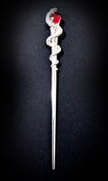 Isolated product shot of the slim silver-tone hair pick against a black background, featuring a scaled snake coiled three times around the upper shaft and a faceted red heart stone held at its head.