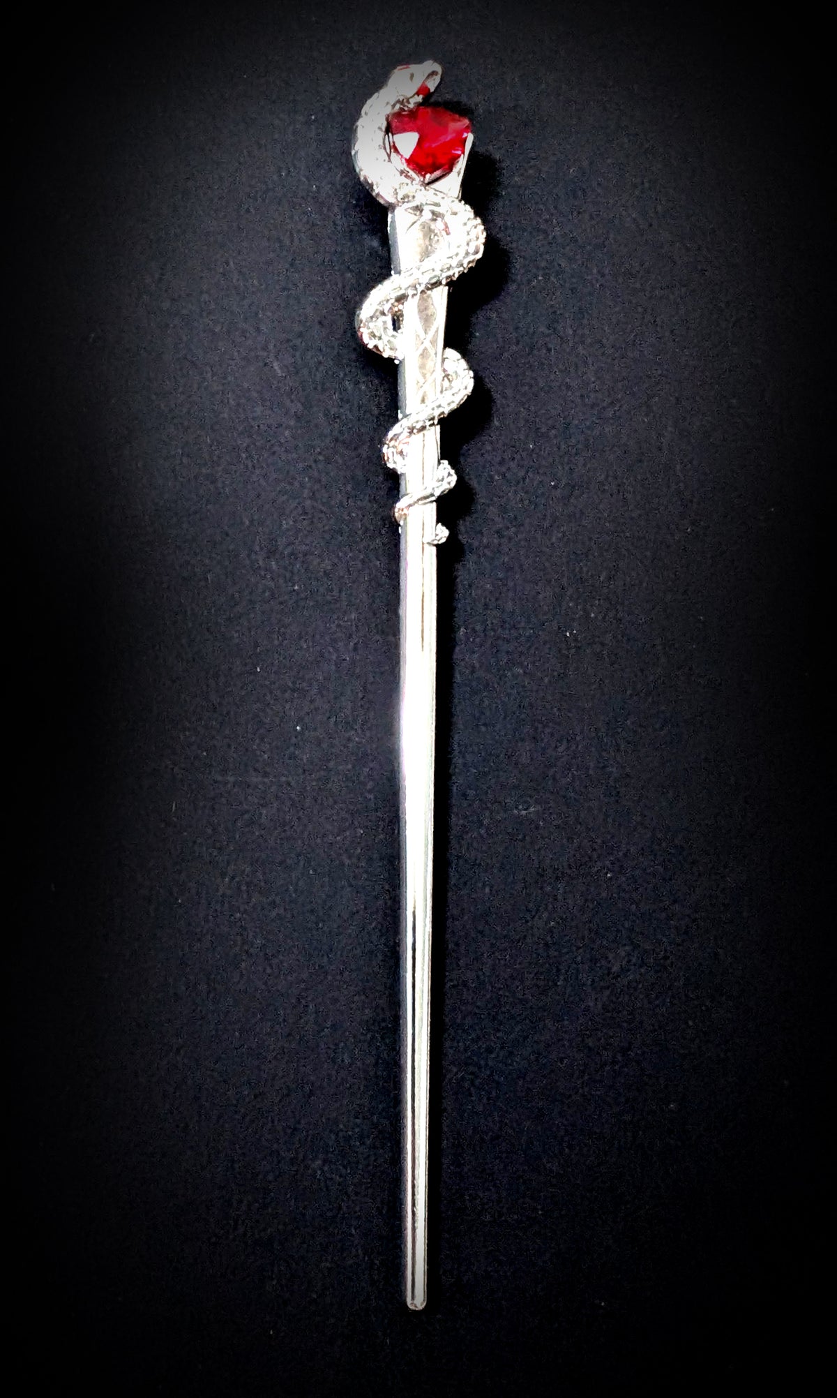 Isolated product shot of the slim silver-tone hair pick against a black background, featuring a scaled snake coiled three times around the upper shaft and a faceted red heart stone held at its head.