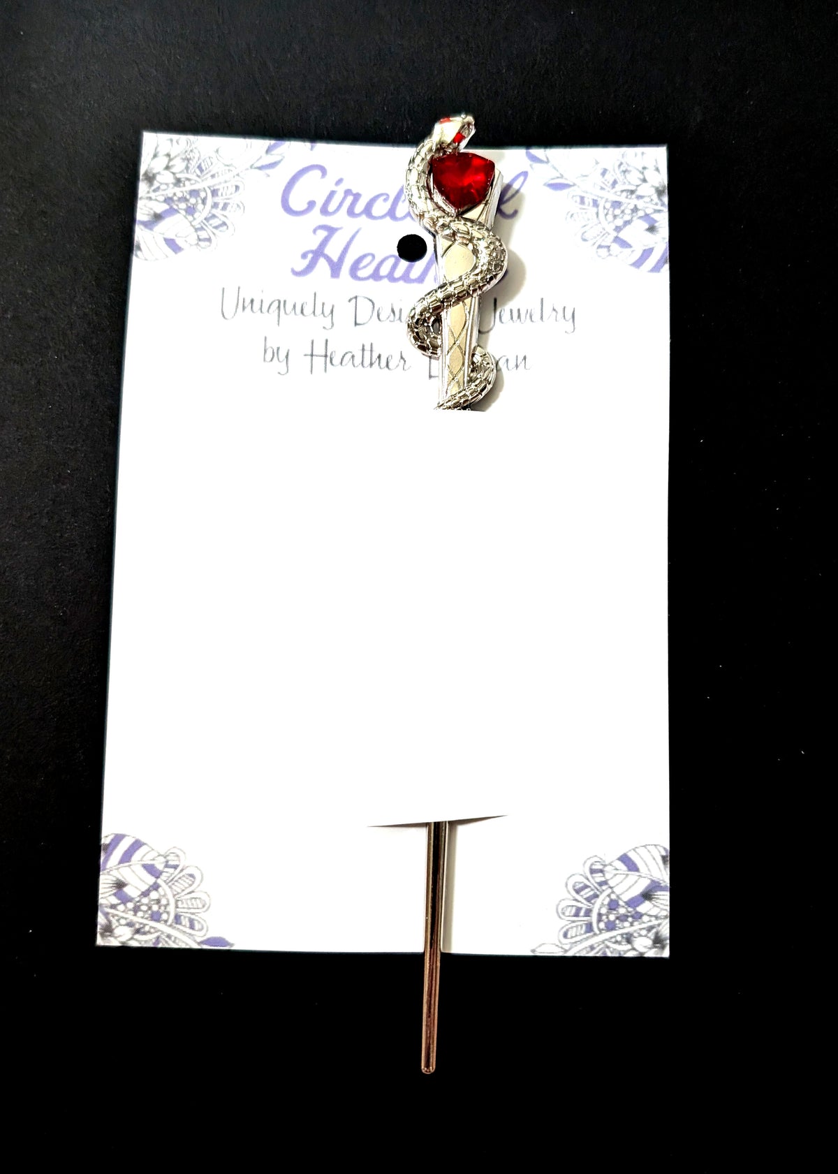 The silver snake hair pick attached to a white backing card featuring a decorative purple mandala pattern and text that reads "Circle of Heather, Uniquely Designed Jewelry by Heather Duggan."