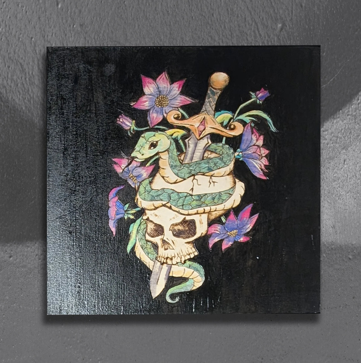 Another view of Serenum Serpentis by CamiThunderInk, an original pyrography, watercolor, and acrylic painting on wood showing an emerald green serpent coiled around a skull pierced by a dagger, with pink and purple flowers on a black background