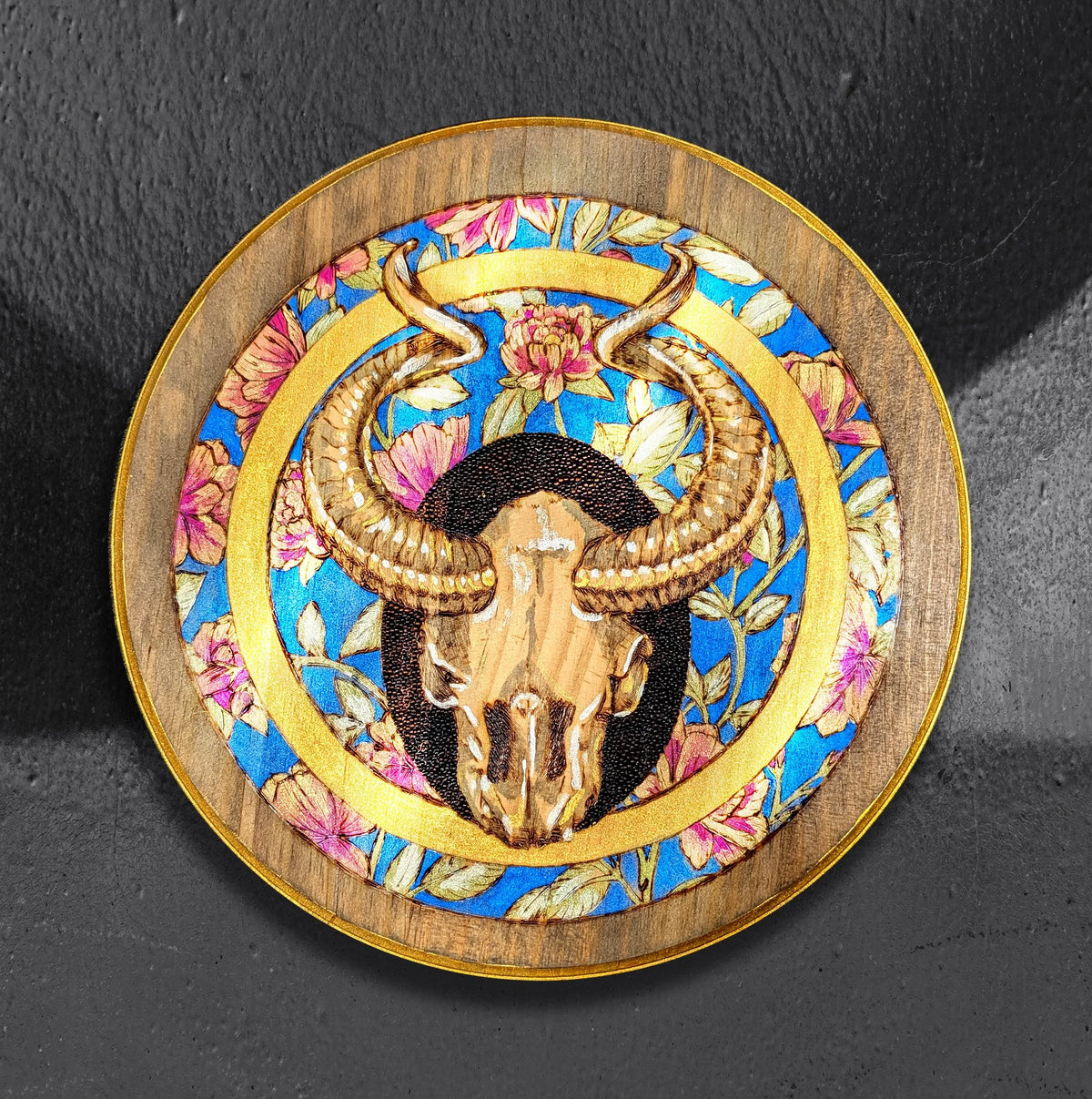 Shimmering Goat Skull by CamiThunderInk, an original pyrography and metallic watercolor painting on a 12 inch pine round of a horned goat skull inside a ring of peonies, with a gold border and stippled black halo