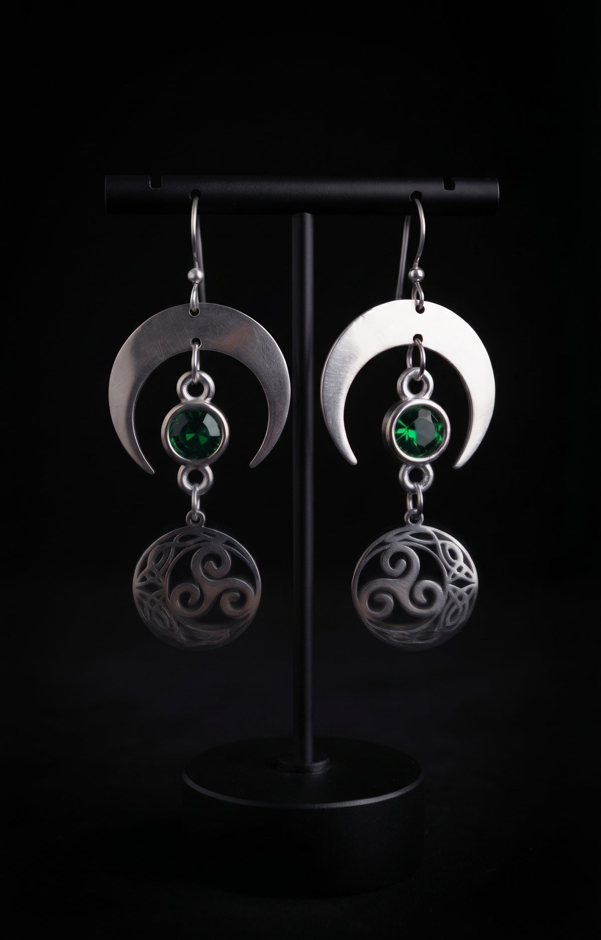 Shimmer Lunar Nocturne Earrings by Circle of Heather, crescent moons with green faceted stones and openwork triple spiral medallions, hanging on a black T-bar jewelry stand.