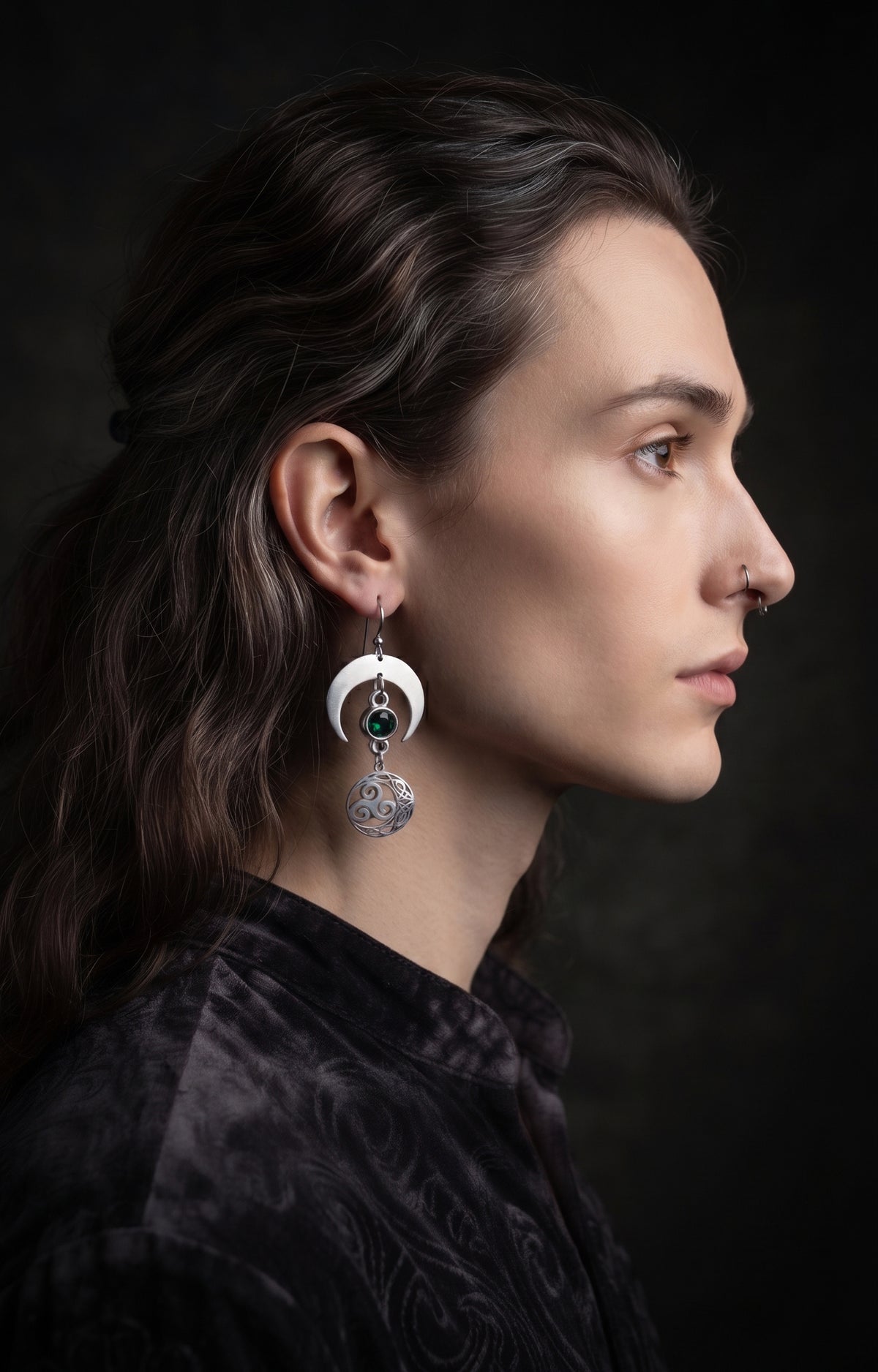 Shimmer Lunar Nocturne Earrings by Circle of Heather worn by a model with long wavy brown hair pulled back and silver nose rings, silver crescent with a green stone above a triple spiral medallion, shown with a dark patterned velvet shirt.