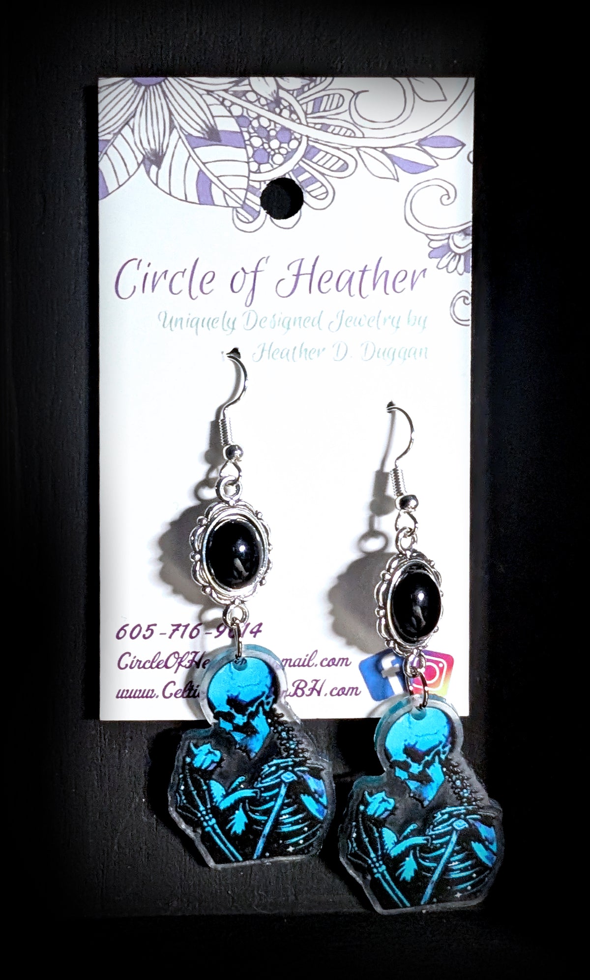 Skele Kitty Snuggles Earrings by Circle of Heather worn by a model with a dark wavy bob and blue glasses, black cabochon above a blue and black printed charm of a skeleton holding a cat.