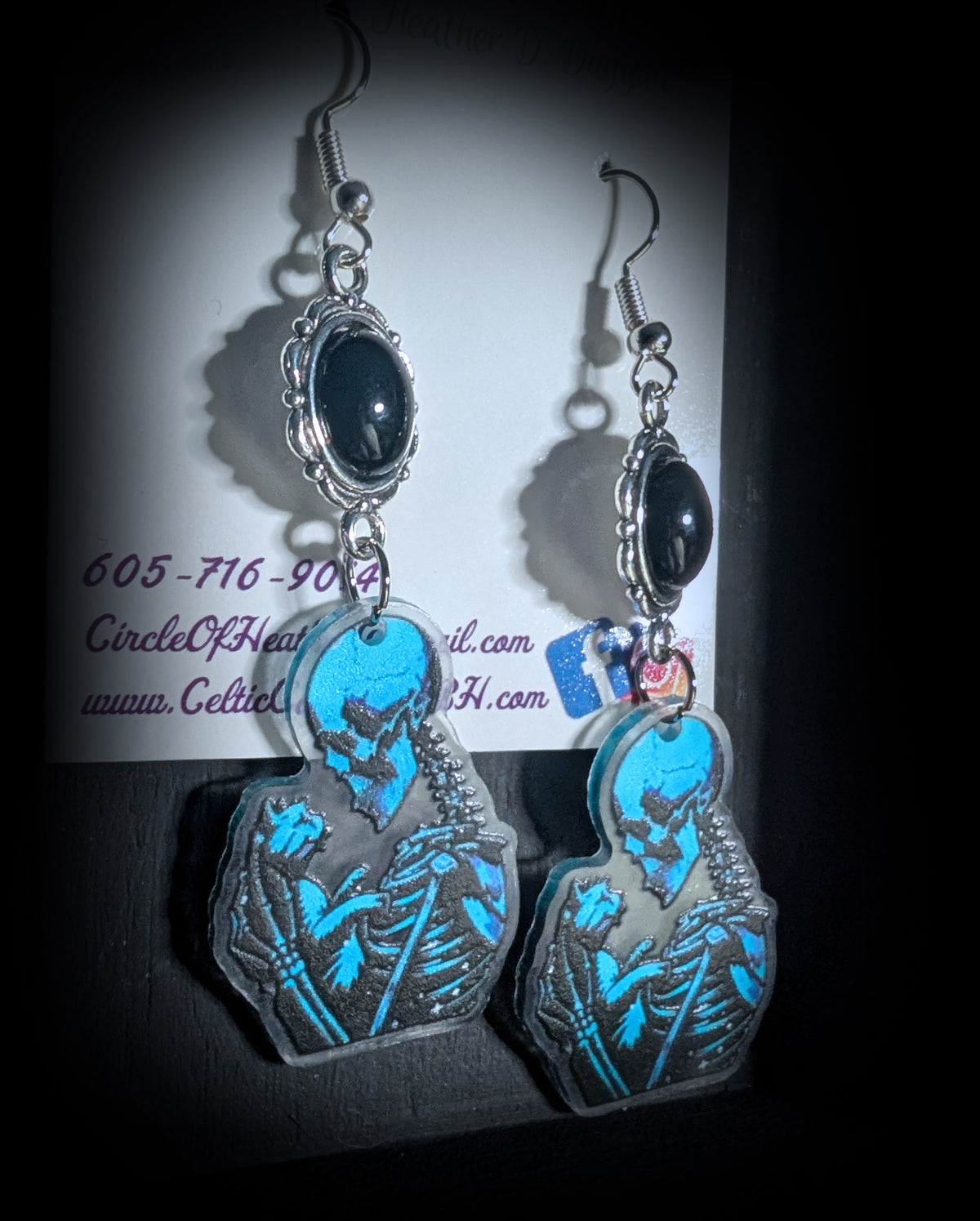 Skele Kitty Snuggles Earrings by Circle of Heather, black cabochons in ornate silver-tone bezels above blue and black skeleton and cat charms, hanging on a black T-bar jewelry stand.