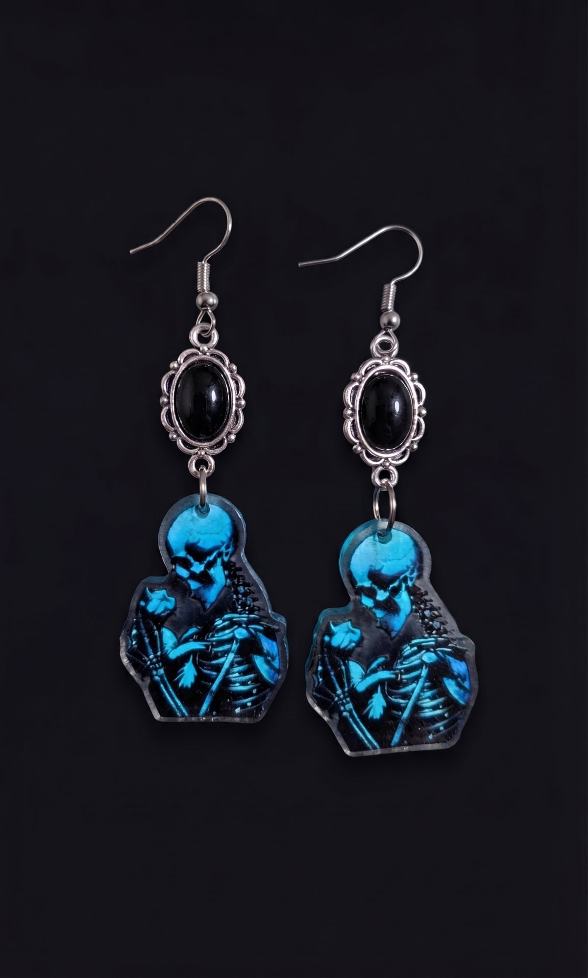 Skele Kitty Snuggles Earrings by Circle of Heather, full view of both earrings showing the black cabochon tops and printed acrylic charms of a skeleton cradling a cat, on a plain black background.