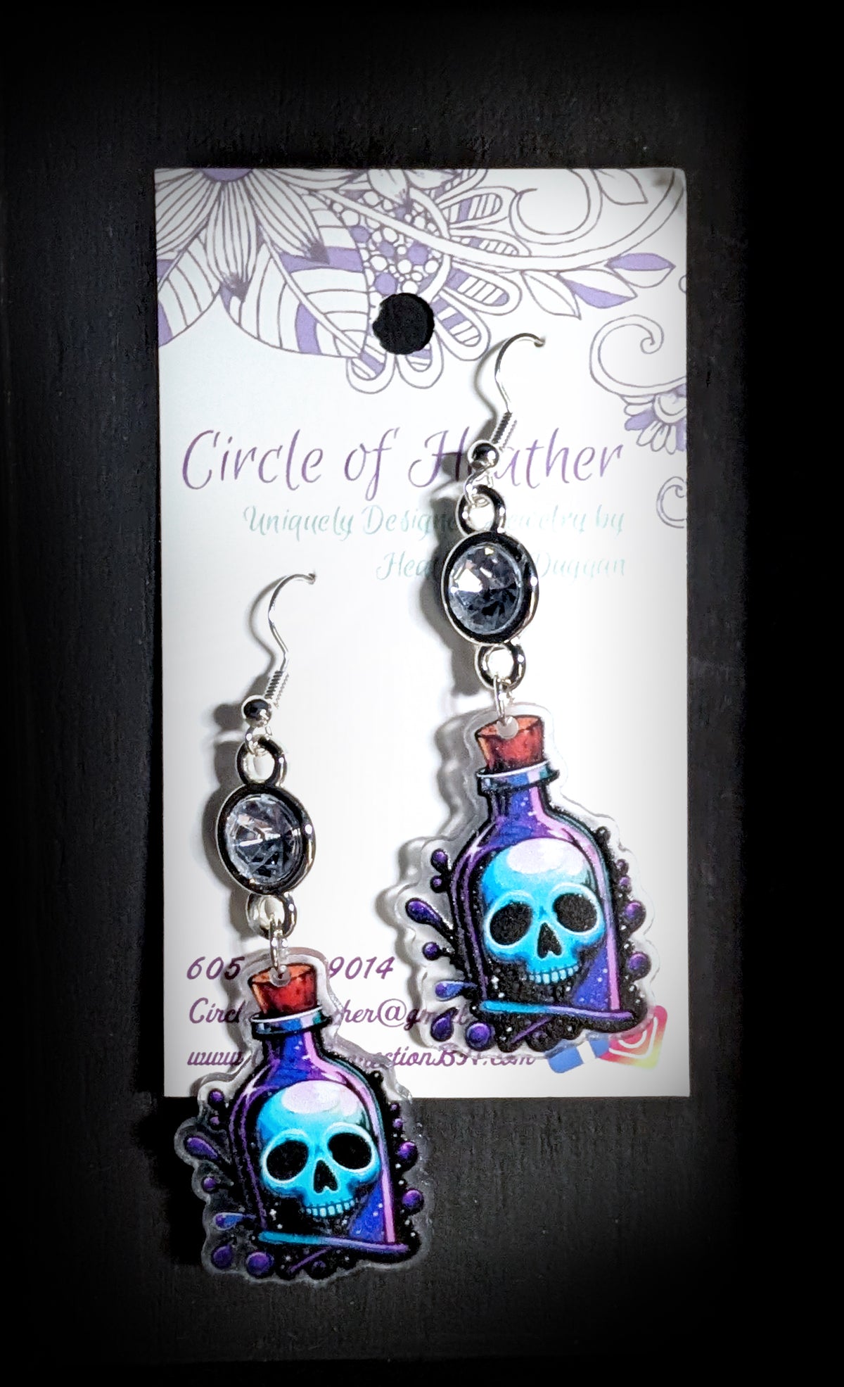 Skull Potion Earrings by Circle of Heather worn by a model with dark hair and teal streaks, clear rhinestone above a printed blue and purple potion bottle charm with a skull inside, against a dark studio background.