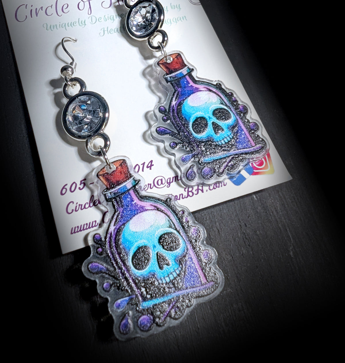 Skull Potion Earrings by Circle of Heather, clear rhinestones in silver-tone bezels above printed acrylic potion bottles with blue skulls and red cork stoppers, hanging on a black T-bar jewelry stand.