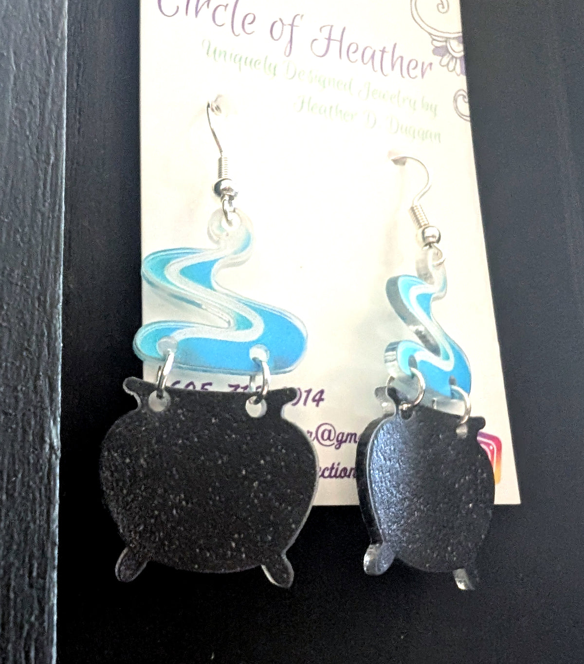 Smoking Cauldron Earrings by Circle of Heather worn by a model with layered gray and blue hair, black glitter cauldron charm with blue and white swirled smoke topper hanging beside a green velvet robe and crystal necklace.