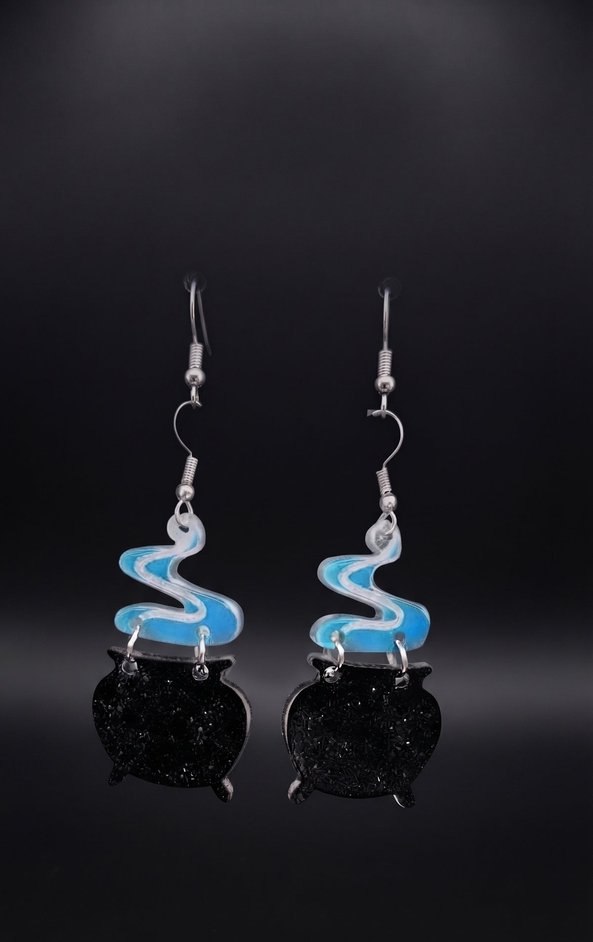 Smoking Cauldron Earrings by Circle of Heather, black glitter cauldron charms with blue and white smoke toppers and silver-tone earwires, hanging on a black T-bar jewelry stand against a dark background with blue backlight.