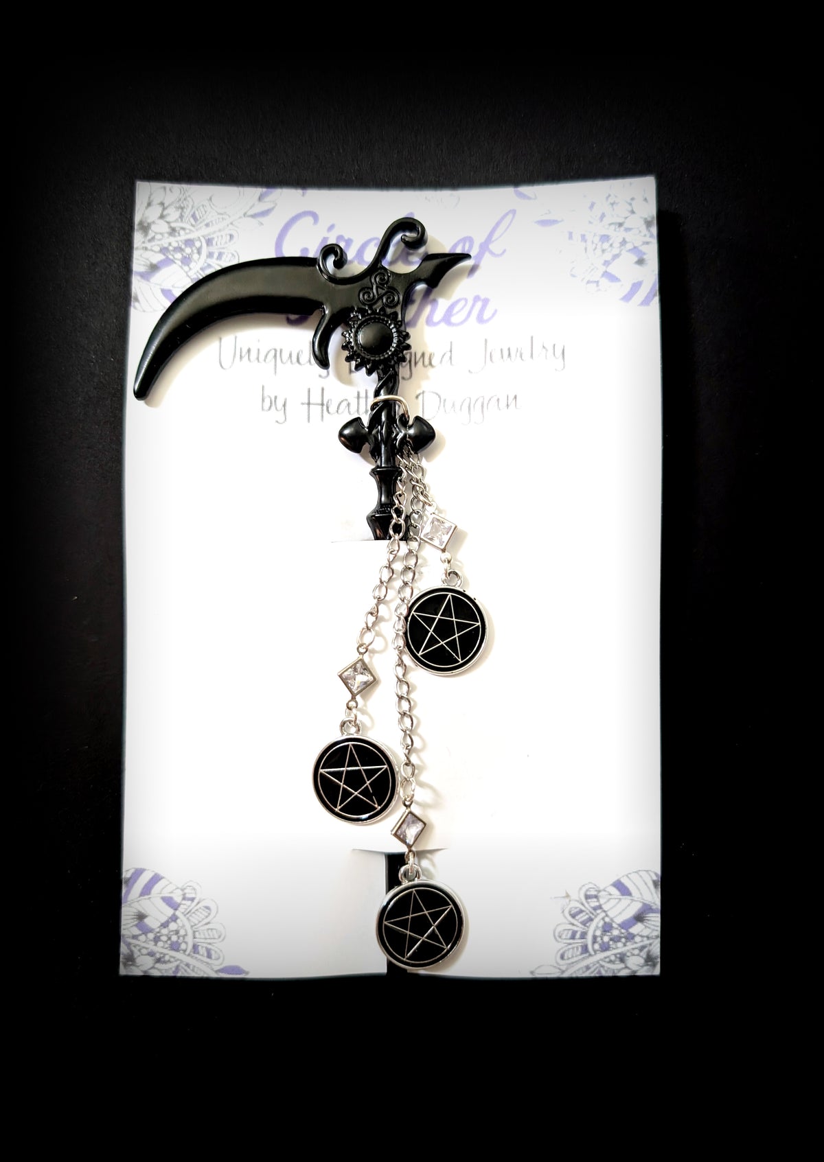 Product shot of the black scythe hair pick against a dark background. It is attached to its white backing card which features a decorative purple mandala pattern and the text "Circle of Heather, Uniquely Designed Jewelry by Heather Duggan." The dangling charms are clearly visible.