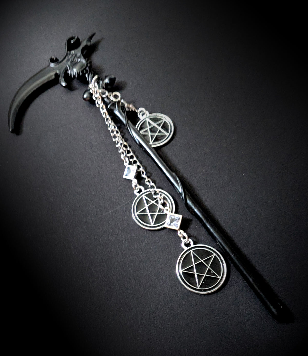 An angled, close-up product shot of the black scythe hair pick against a dark background. It highlights the curve of the glossy black blade, the black cabochon setting, and the three dangling silver pentacle charms connected by rhinestone links.