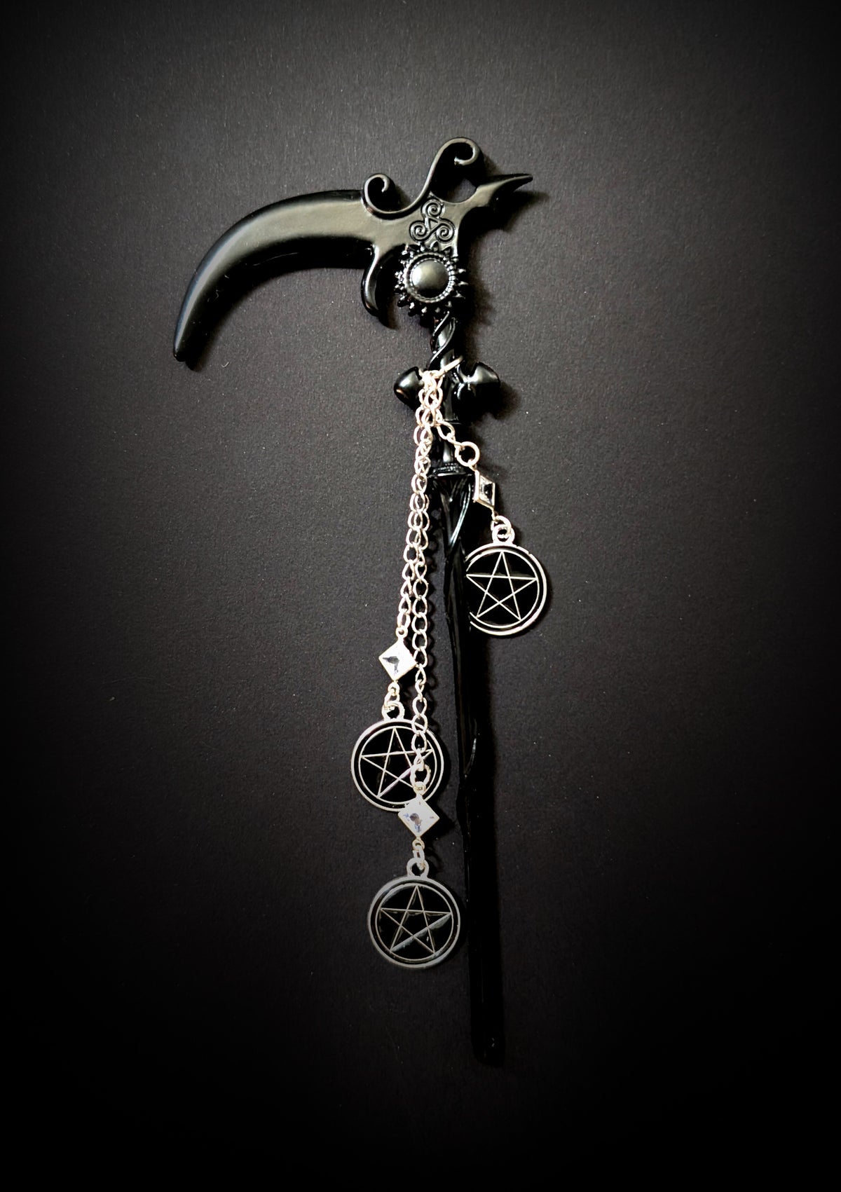 Close-up product shot of the black scythe-shaped hair pick against a dark background. It features ornate scrollwork and a triple spiral detail at the neck. A round black cabochon is set in a sunburst bezel at the joint. Three silver pentacle charms on staggered chains with clear rhinestone links hang from the neck.