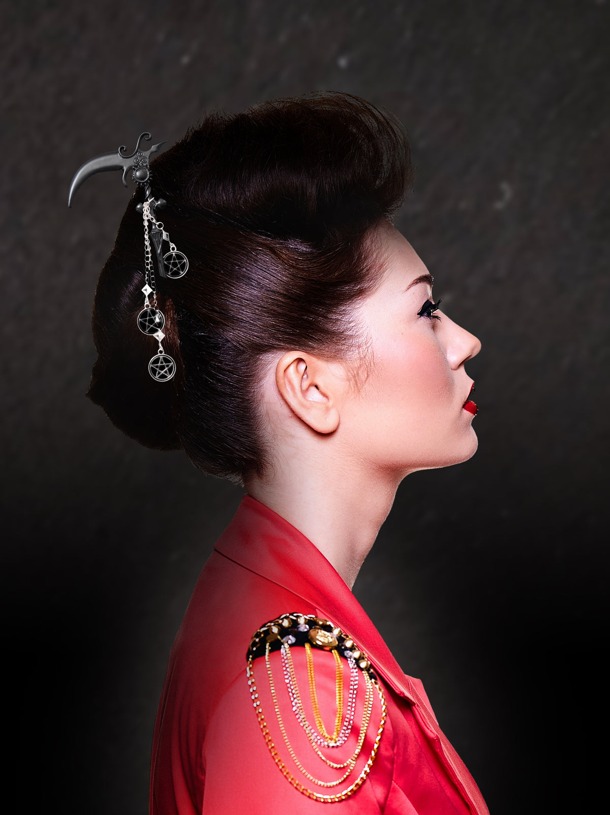 A side profile of a model with dark hair styled in an elaborate dark updo, pierced by the long black scythe hair pick. Three silver pentacle charms dangle from fine chains linked with clear square rhinestones. The model wears a red jacket with chain epaulets against a dark, textured background.