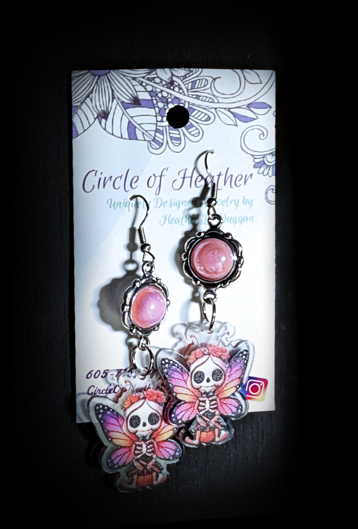 Sweet Pink Skull Fairy Earrings by Circle of Heather worn by a model with a purple and blue pixie cut, pale pink cabochon above a printed skull fairy charm with pink and orange butterfly wings, against a gray wood background.