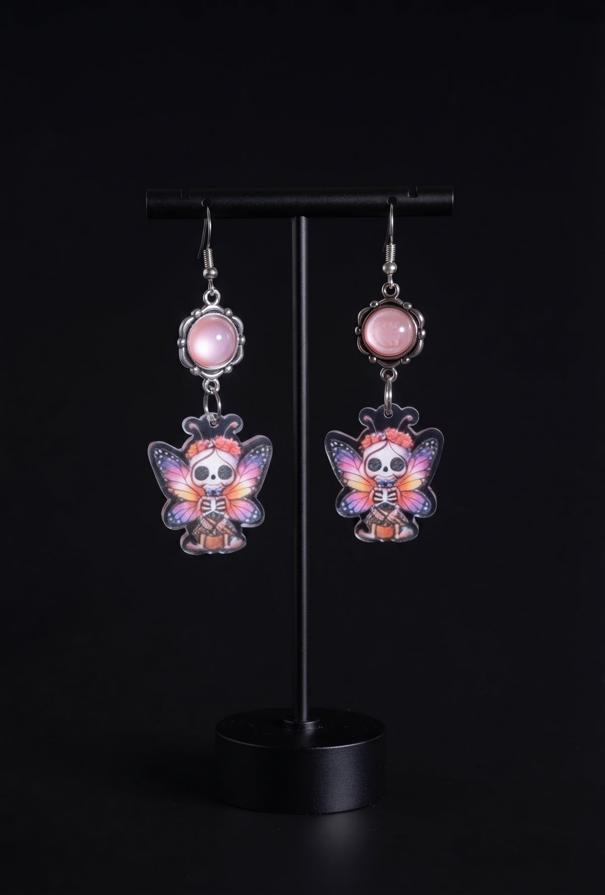 Sweet Pink Skull Fairy Earrings by Circle of Heather, pink cabochons in ornate silver-tone bezels above printed acrylic skull fairy charms with coral flower crowns, hanging on a black T-bar jewelry stand against a black background.
