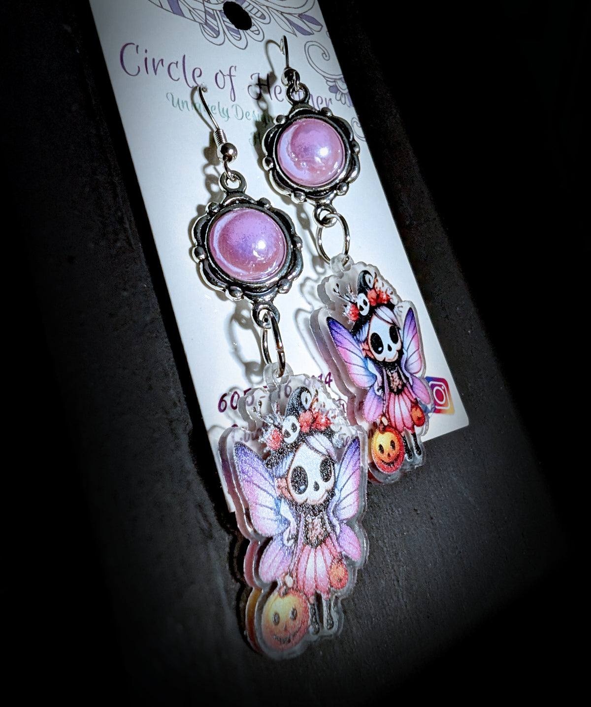 Treat Skull Fairy Earrings by Circle of Heather, angled close-up showing the scalloped bezel around the pink cabochon and the witch hat and wing detail on the printed charms, shown on the branded Circle of Heather card.