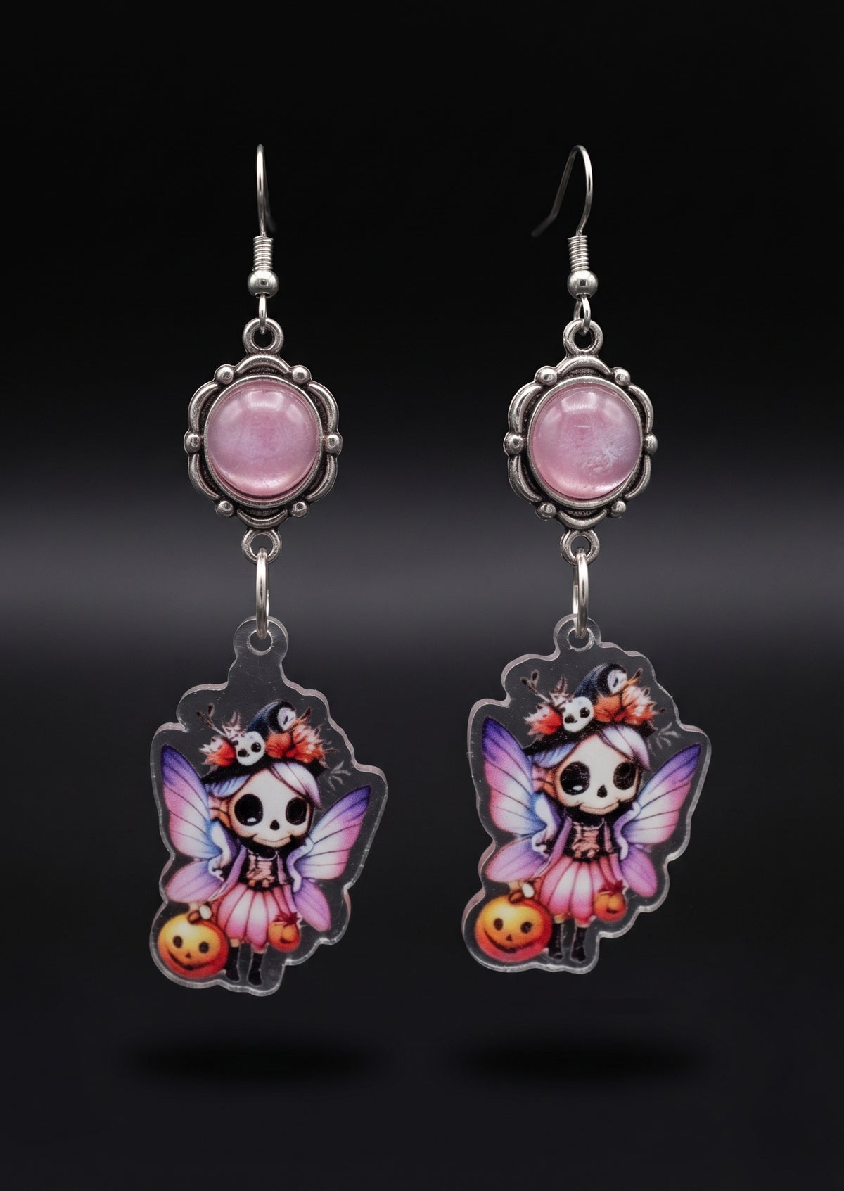 Treat Skull Fairy Earrings by Circle of Heather, full view of both earrings showing the pink cabochon tops and skull fairy charms with pink and purple wings, pink tutus and jack-o-lanterns, on a plain black background.
