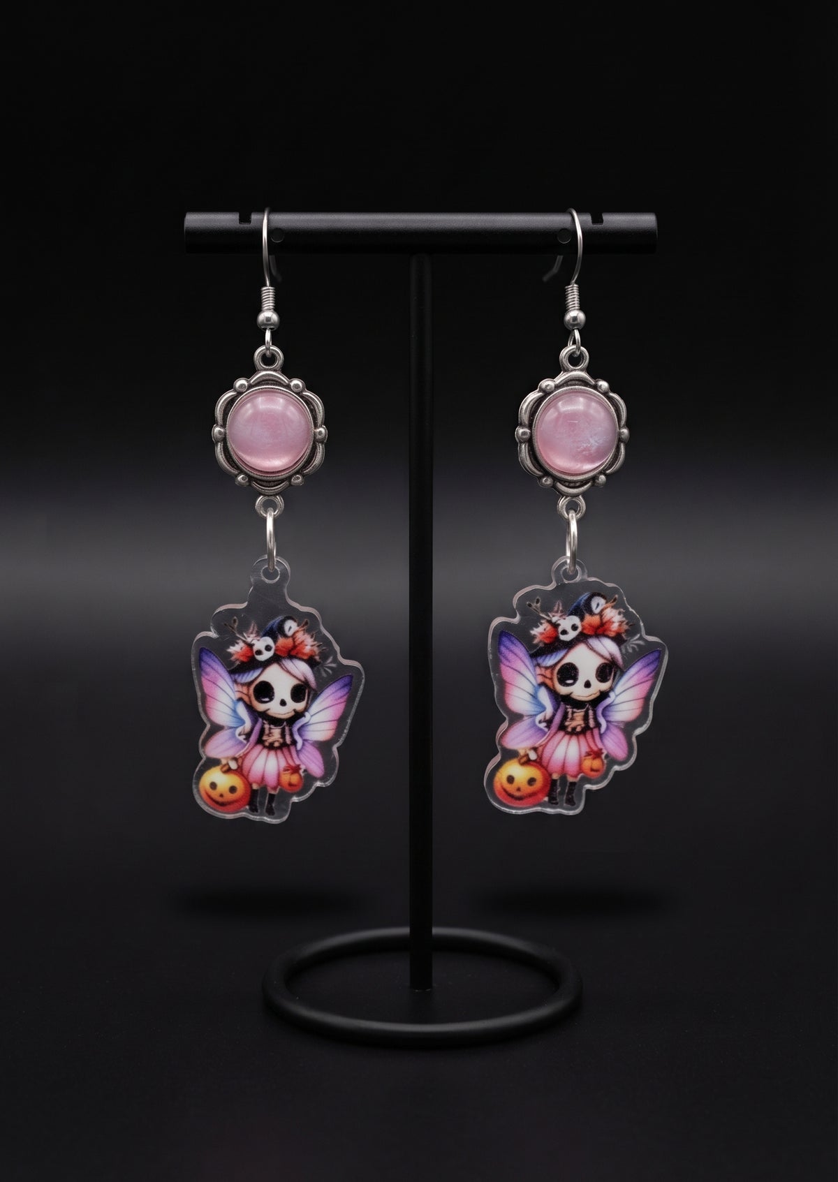 Treat Skull Fairy Earrings by Circle of Heather, pink cabochon tops and printed skull fairy charms hanging on a black T-bar jewelry stand against a black background.