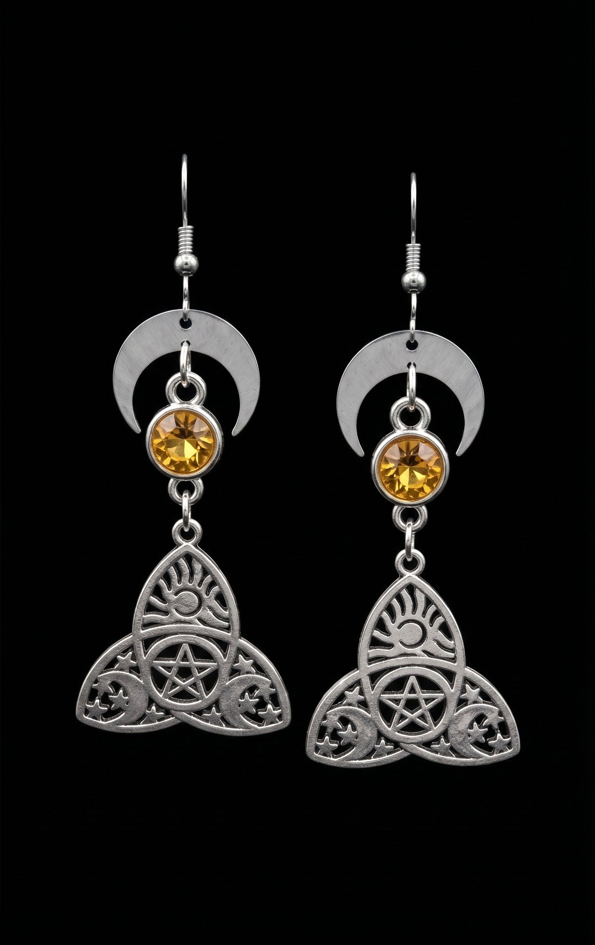 Trinity Lunar Glow Earrings by Circle of Heather, full view of both earrings showing the crescent moon, faceted amber stone and trinity knot charm with pentacle, sun and moon cutwork, on a plain black background.
