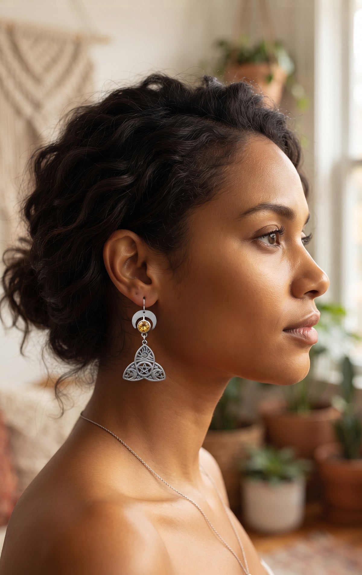 Trinity Lunar Glow Earrings by Circle of Heather worn by a model with dark curly hair in an updo, silver crescent moon above an amber stone and a trinity knot charm with a pentacle center, in a sunlit room with plants and macrame.