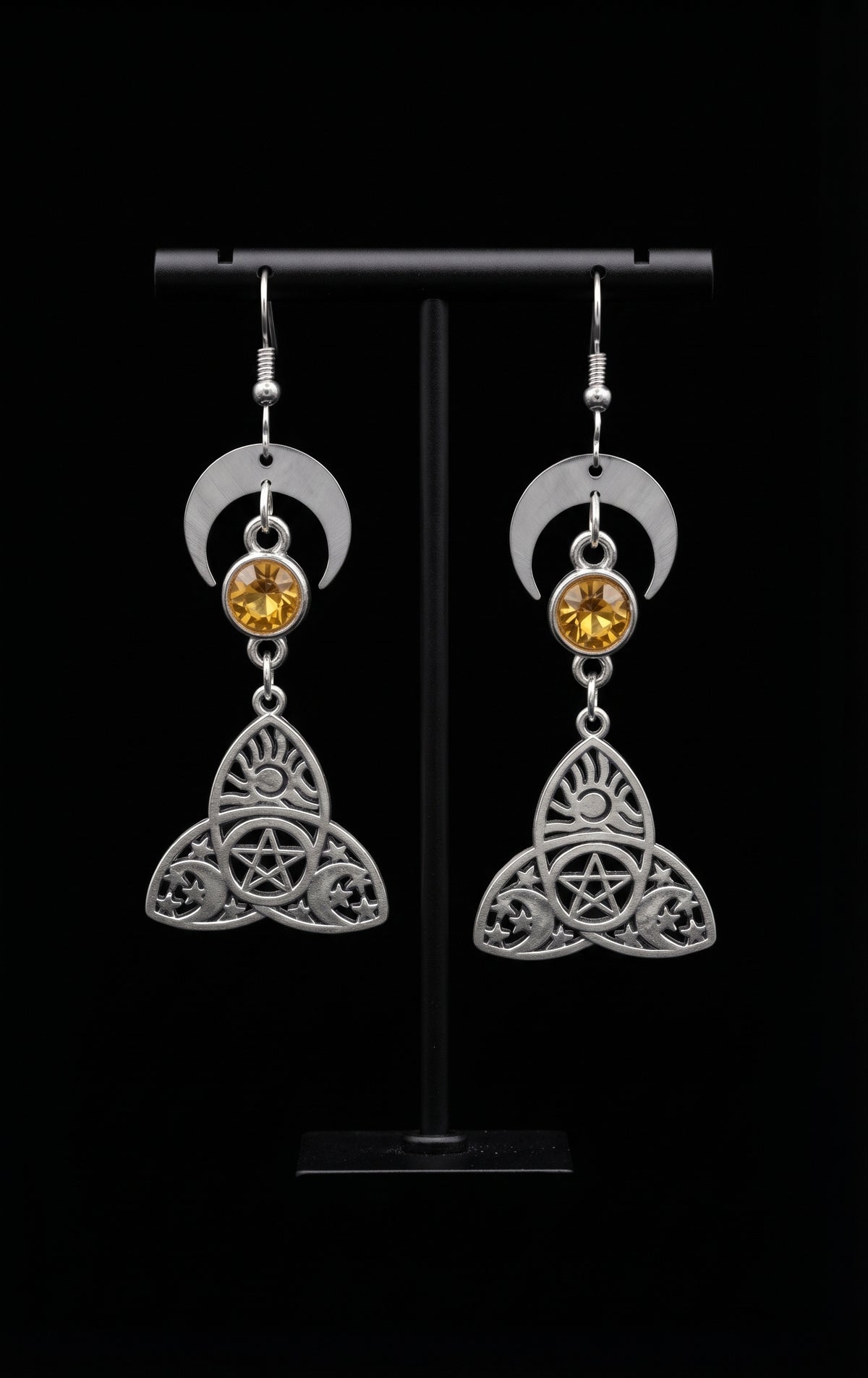 Trinity Lunar Glow Earrings by Circle of Heather, crescent moon tops with amber stones and trinity knot pentacle charms, hanging on a black T-bar jewelry stand against a black background.