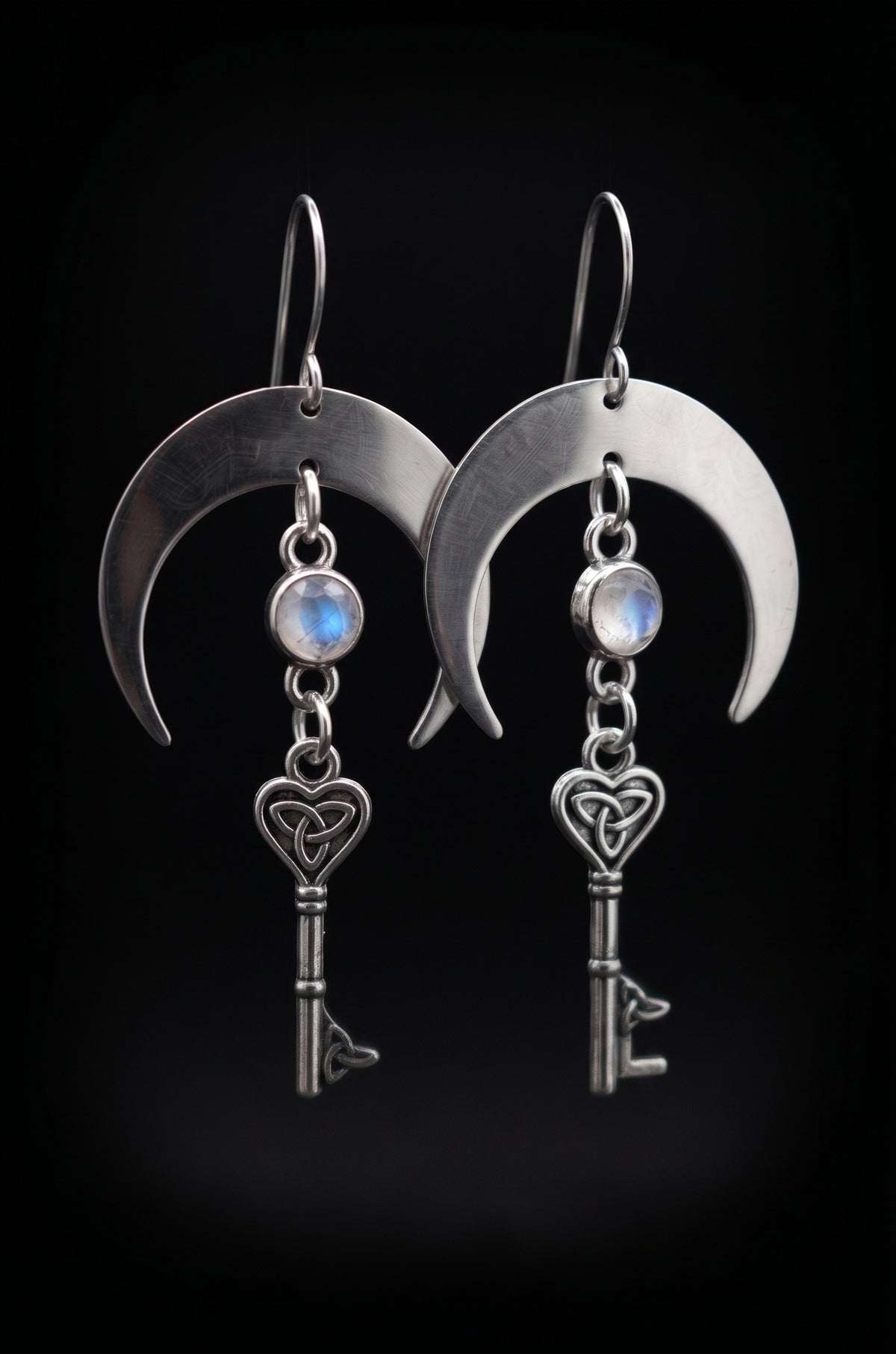 Unlock the Moon Earrings by Circle of Heather, full view of both earrings showing the brushed crescent moons, blue flash in the opalescent stones and the heart-topped skeleton keys, on a plain black background.