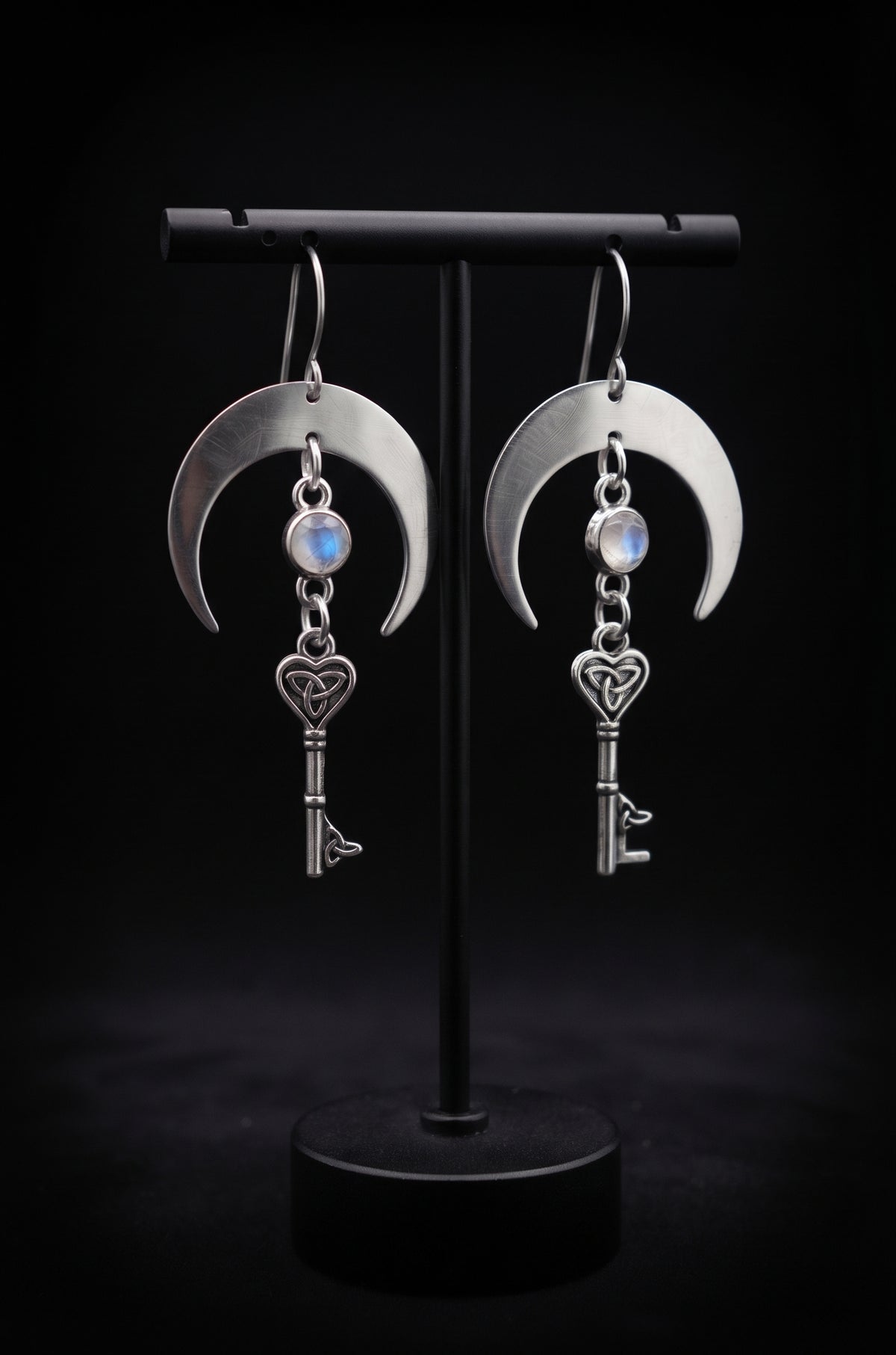 Unlock the Moon Earrings by Circle of Heather, wide silver crescents with opalescent stones and Celtic trinity knot key charms, hanging on a black T-bar jewelry stand.