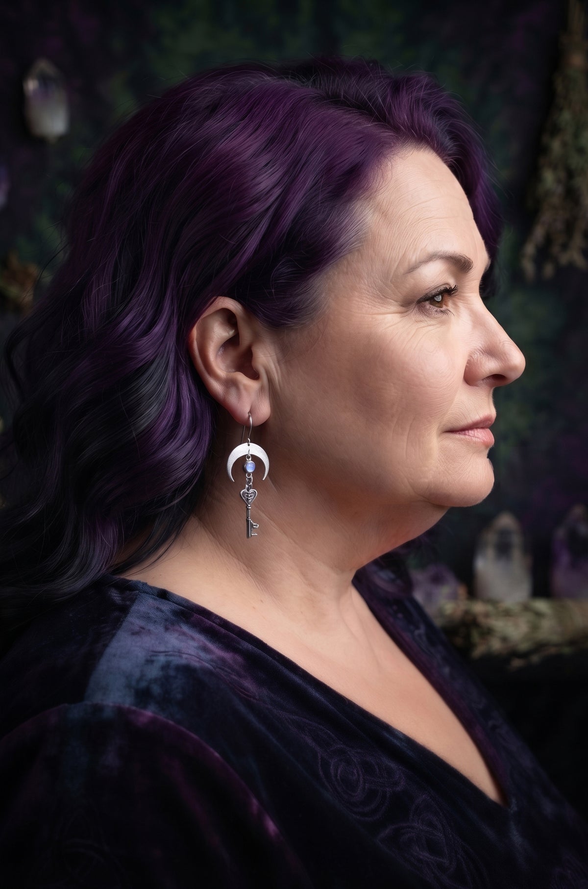 Unlock the Moon Earrings by Circle of Heather worn by a model with deep purple wavy hair, brushed silver crescent with a pale opalescent stone above a Celtic trinity knot key, shown with a dark purple velvet top and crystals behind.