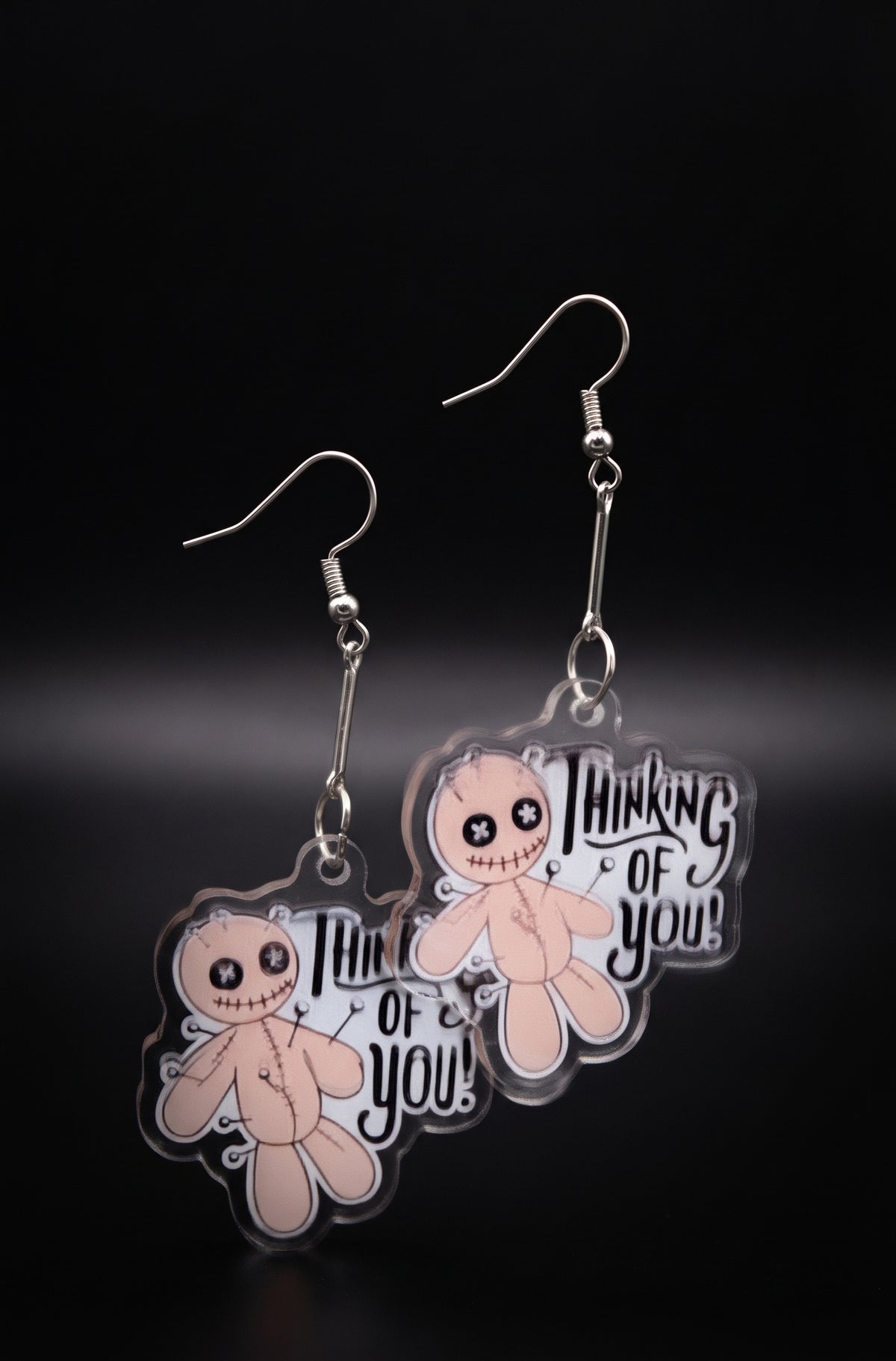 Voodoo Doll Thinking of You Earrings by Circle of Heather, full view of both charms showing the cross-stitch eyes, stitched mouth and hand-lettered "Thinking of you!" text, on a plain black background.